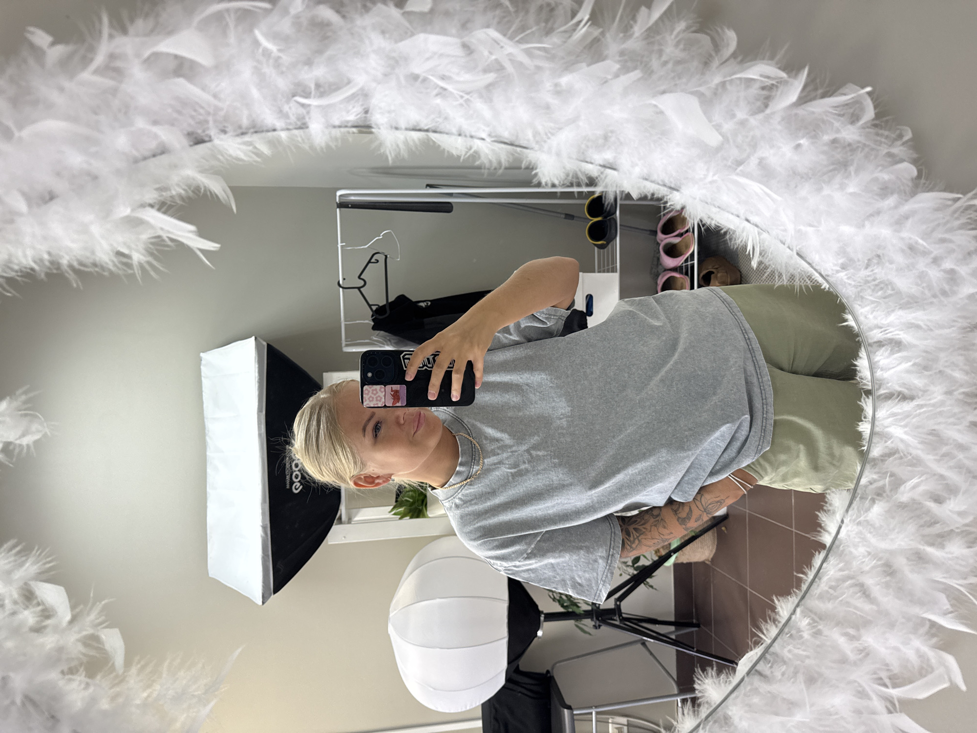 A person is taking a mirror selfie. The mirror is circular and decorated with white feathers around the edge. The person is wearing a gray t-shirt and green pants. In the background, there is a clothing rack with clothes and shoes on it, and two softboxes for lighting. The flooring is brown tile. The location is a room with neutral-colored walls.