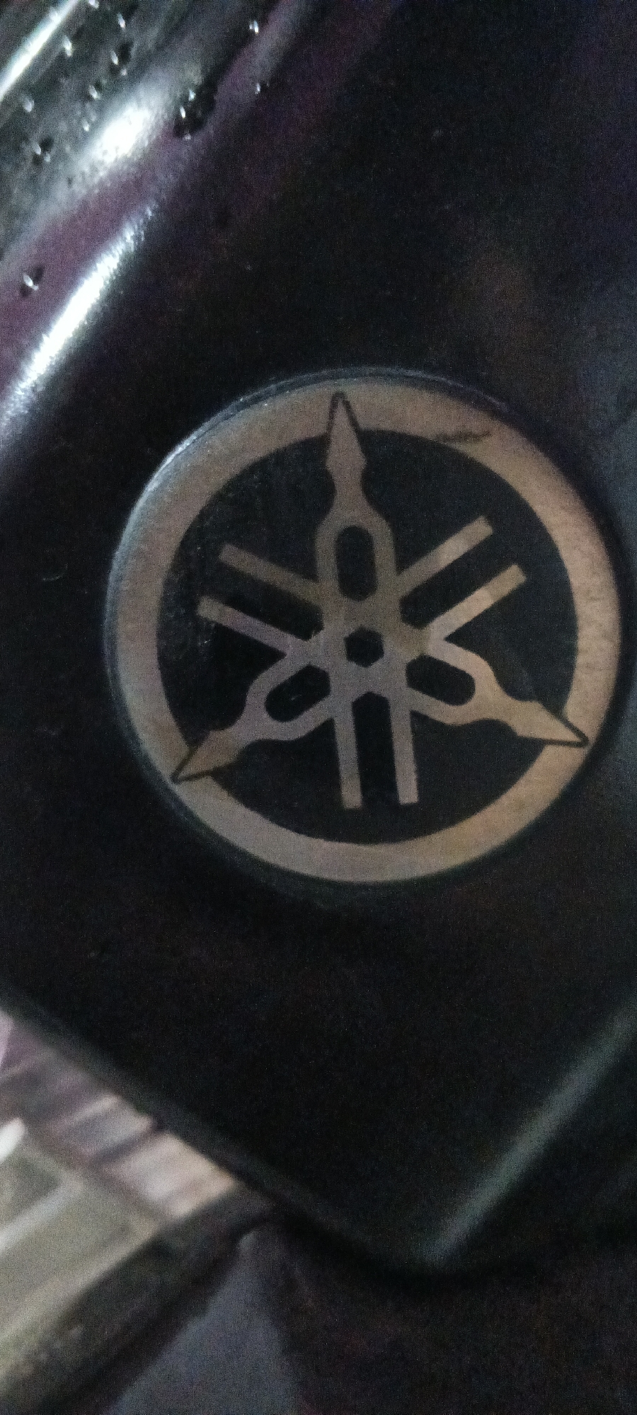 The image shows a close-up of the Yamaha logo on a dark, possibly metallic, surface. The logo is a circular emblem with three interlocking tuning forks radiating from the center, enclosed by a circular border. The logo appears to be silver or a metallic gray against a black background. The surface it's on is slightly reflective and shows some subtle textures or imperfections. The lighting in the image is dim, creating shadows and highlighting the contours of the logo and the surrounding material. There are some small, irregular reflections or water droplets visible on the surface to the left of the logo. No people or specific city locations in Suka Sari, Indonesia are visible in this image.