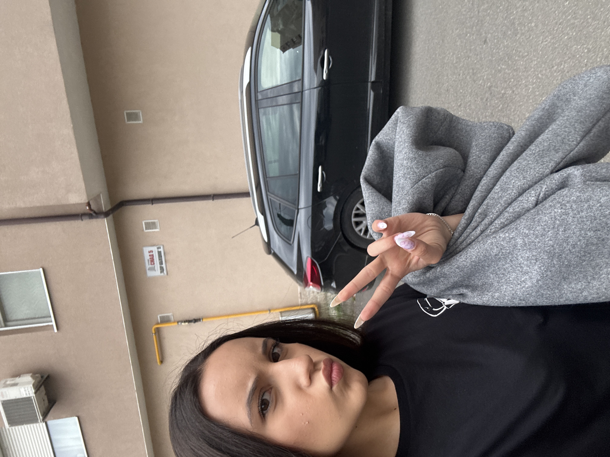 The media file is a vertical, selfie-style photograph of a young woman posing in front of a black station wagon. The woman is in the foreground, looking towards the camera with her lips slightly puckered. She has dark hair and is wearing a black t-shirt. Her right hand is raised, forming a peace sign with her fingers. Her nails are painted with a light pink and purple marble design. She is also wearing a grey sweater or jacket draped over her arm.
In the background, the black station wagon is parked. Part of a beige building with windows and some exterior pipes is visible. One of the pipes appears to be yellow and is attached to the building. There is also a sign on the building with some text, but it is difficult to read clearly. The scene appears to be outdoors, possibly in a residential area or parking lot. The lighting suggests it is daytime, and the weather appears to be neutral. The woman's pose and facial expression convey a casual and playful mood.