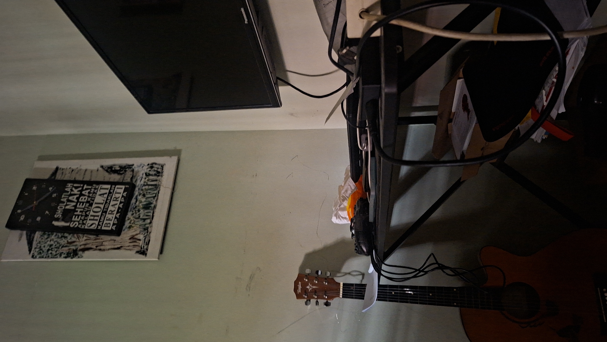 A dimly lit room in Padang, Indonesia, features an acoustic guitar leaning against a wall. A black metal desk is positioned in the foreground, cluttered with electronic devices, wires, and papers. A flat-screen television is mounted on the wall above the desk, reflecting the ambient light. A framed picture hangs on the wall to the left of the television, displaying text and an illustration. The walls are painted a light green and show signs of age. The scene appears to be a personal space, possibly a home office or entertainment area.