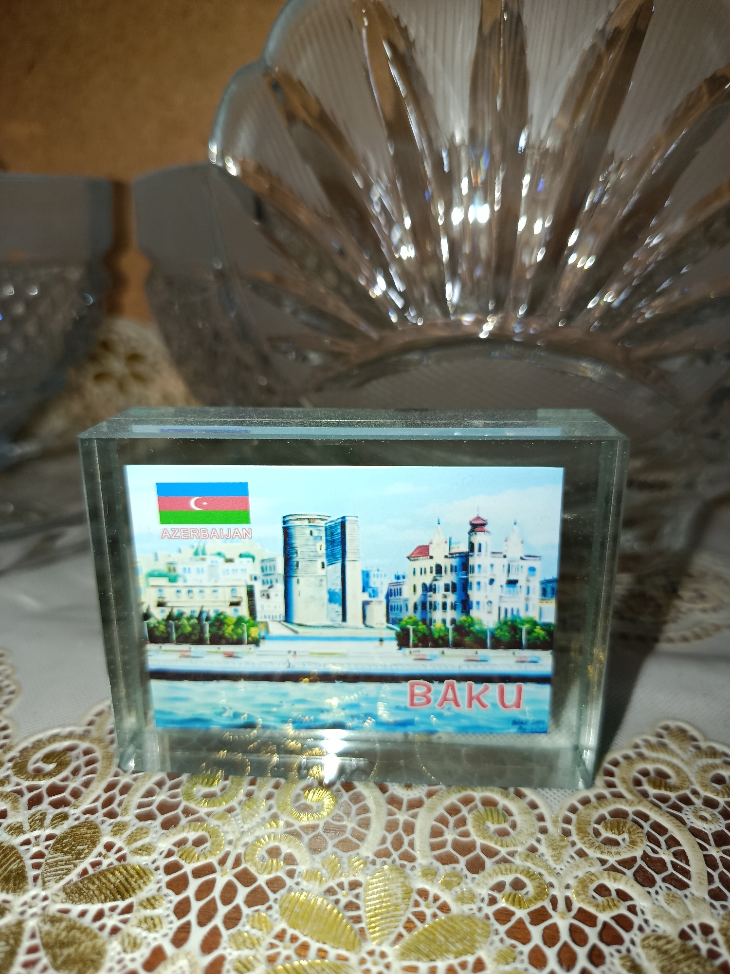 A close-up shot focuses on a glass paperweight displaying a picture of the city of Baku, Azerbaijan. The image within the paperweight features the Azerbaijan flag, the word "AZERBAIJAN" in white text, a waterfront scene with buildings, and the word "BAKU" in pink text. The paperweight rests on a white and gold embroidered doily. In the background, out of focus, are parts of clear glass bowls or vases. The lighting creates reflections and bokeh effects on the glass objects and the doily. There are no people visible in the image.