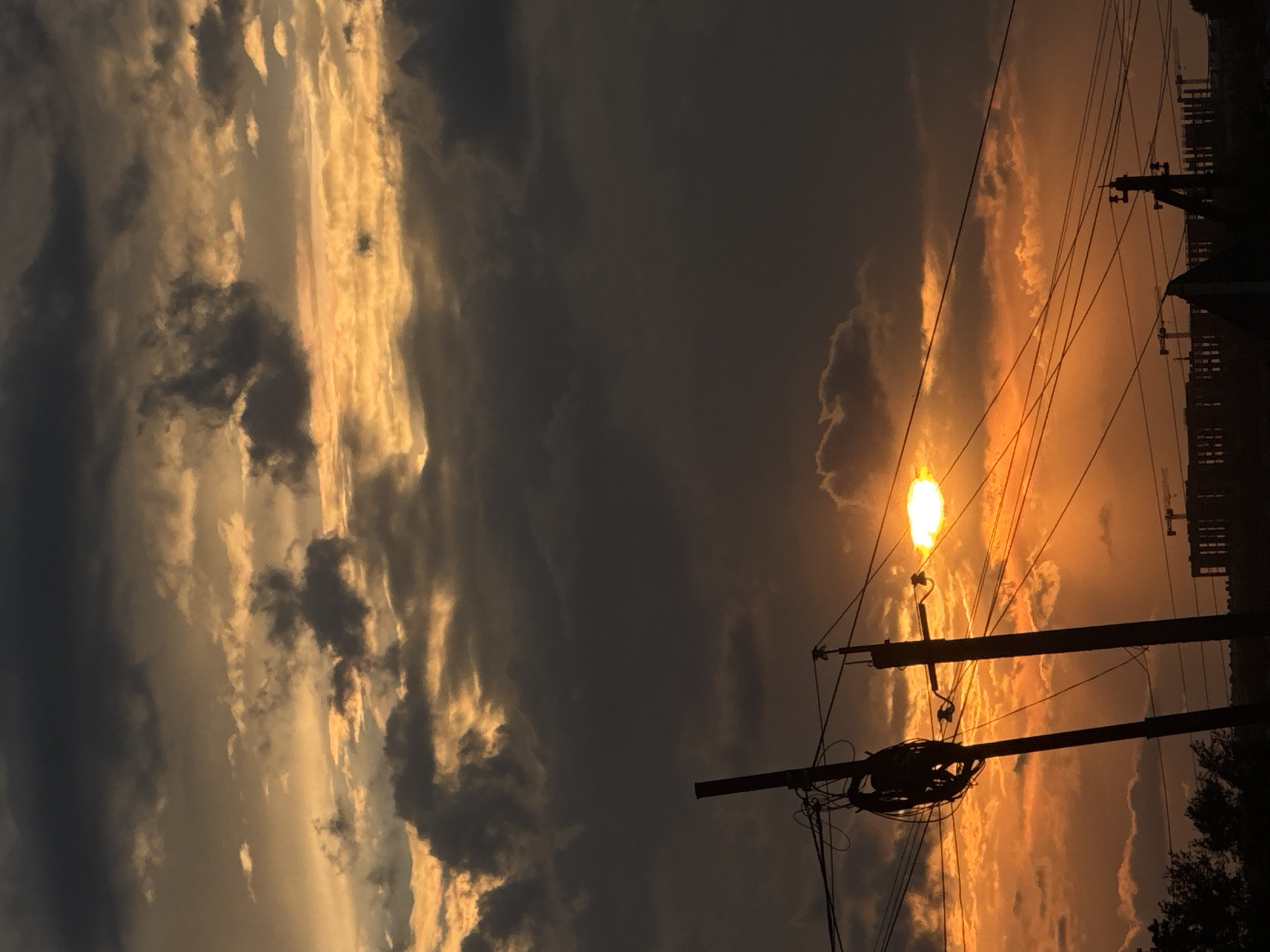 This media file captures a dramatic sunset over Mikhaylovsk, Russia. The scene is dominated by a sky filled with a mix of dark, brooding storm clouds and streaks of bright, golden-orange sunlight. The sun itself is a brilliant, glowing orb peeking through the clouds, casting an intense orange hue across the sky and illuminating the edges of the clouds.
In the foreground, silhouetted against the vibrant sky, are power lines and utility poles. The lines crisscross the frame, with one pole notably topped with a bright, incandescent light, adding to the surreal glow of the sunset. The intricate network of wires suggests an urban or semi-urban environment. In the far background, dark, indistinct shapes suggest buildings or structures, further anchoring the location within a populated area.
The time of day is clearly sunset, given the intense colors and the position of the sun. The weather appears to be a transition between a storm and clear skies, with the dark clouds hinting at recent or impending rain, while the bright sun indicates a break in the weather. The overall mood is one of dramatic beauty and a touch of foreboding due to the storm clouds, juxtaposed with the warmth and intensity of the setting sun. There is no visible text in the media file.