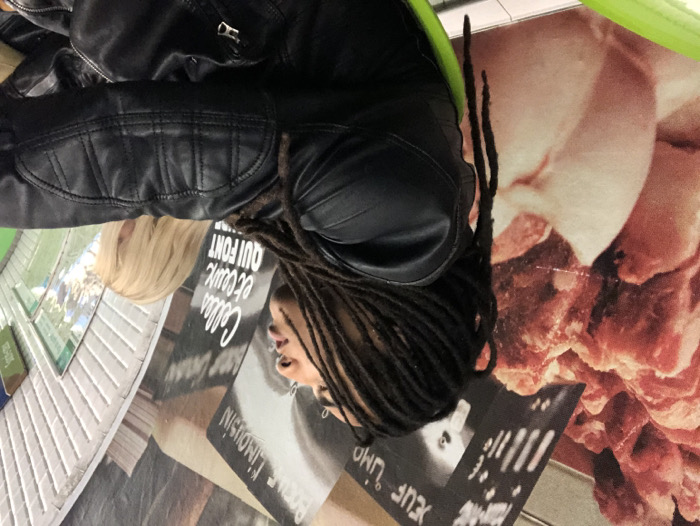 The scene is set within a Paris Metro station, indicated by the distinctive curved, ribbed architectural features and French language advertising. In the foreground, a person with dark skin and long dreadlocks is seated, facing right and looking slightly upwards with a neutral expression. They are wearing a black leather jacket with quilted detailing on the shoulders and arms. A bright green element, likely part of a seat, is visible beneath them on the lower left. Partially obscured behind and to the right of the primary subject, a second individual with long blonde hair is visible from the back, also wearing a black jacket. The background wall features a large advertisement depicting cuts of raw red meat, with visible French text including "BOEUF LIMOUSIN" (Limousin Beef) and "CELLES ET CEUX QUI FONT" (Those who make/do). The architecture to the far right displays a characteristic white, ribbed, curved ceiling structure of the Metro. No direct interaction occurs between the two visible individuals.