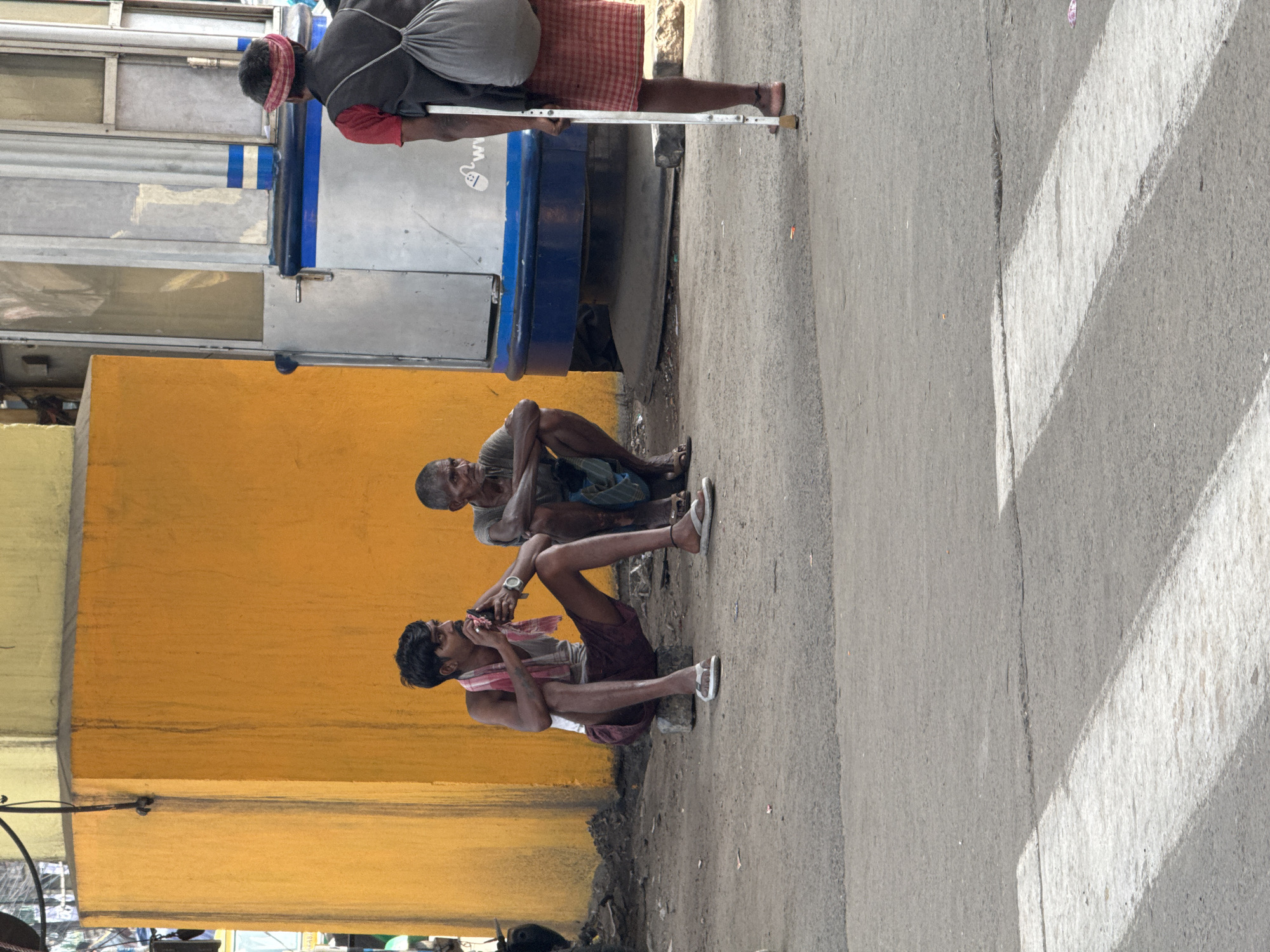 The image depicts a street scene in Kolkata, India. Two men are sitting on the pavement near a yellow wall, one using a mobile phone. Another man with a crutch walks beside them. There is a bus or commercial vehicle next to them. The road is gray, and the weather is bright.