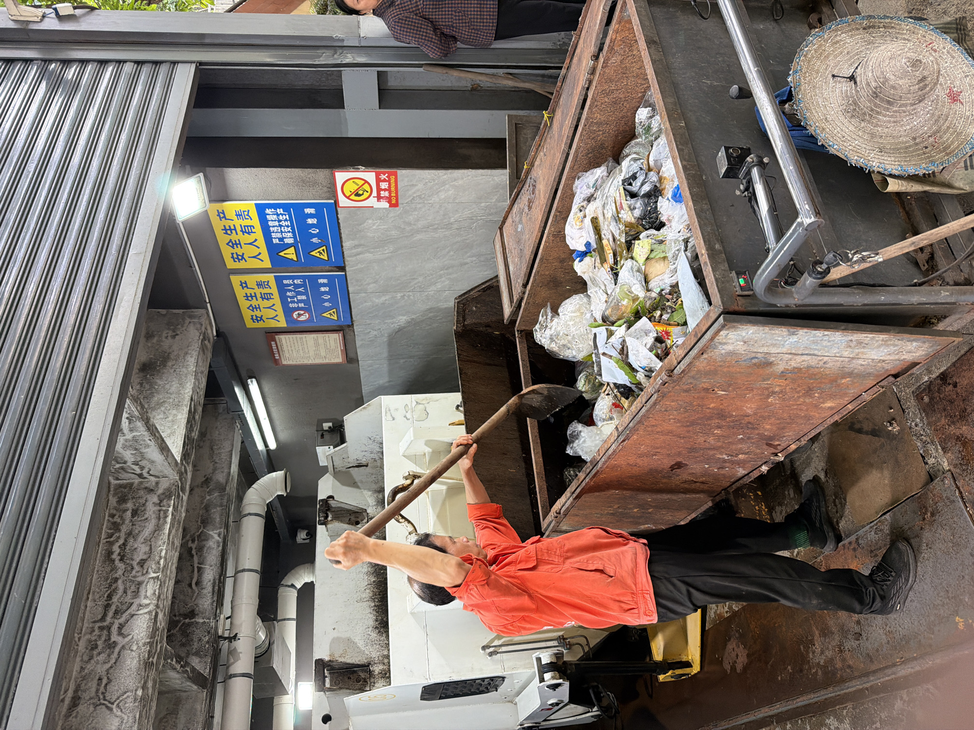 In Yuzhong District, China, a man in an orange shirt and black pants uses a large shovel to move trash into a rusty, open-top dumpster. The dumpster is filled with plastic bags and various types of refuse. Behind him, a large white garbage truck is partially visible. On the wall behind the dumpster, there are several safety signs in Chinese. One sign indicates "No Smoking," and others display safety regulations. To the right of the dumpster, a woman in a plaid jacket stands near the entrance. The scene appears to be a waste disposal or collection point.