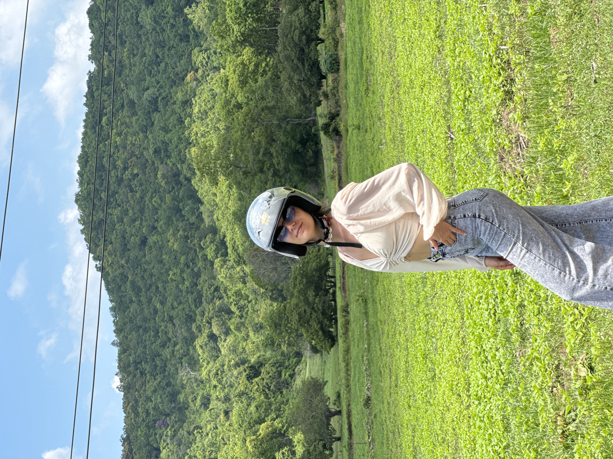 A young woman stands in a bright, green field, a lush, tree-covered mountain range rising behind her under a clear blue sky with scattered clouds. The woman, wearing a white, ruched crop top, light-wash wide-leg jeans, sunglasses, and a silver motorcycle helmet, is positioned in the center of the frame, looking directly at the camera. Her right hand is casually tucked into her jeans pocket. Power lines are visible stretched across the top of the image, above the mountain. The field is densely covered with low-lying green plants, with scattered larger trees in the mid-ground. This scene appears to be in Ham Thuan Bac, Vietnam, a region known for its natural landscapes and agricultural fields.