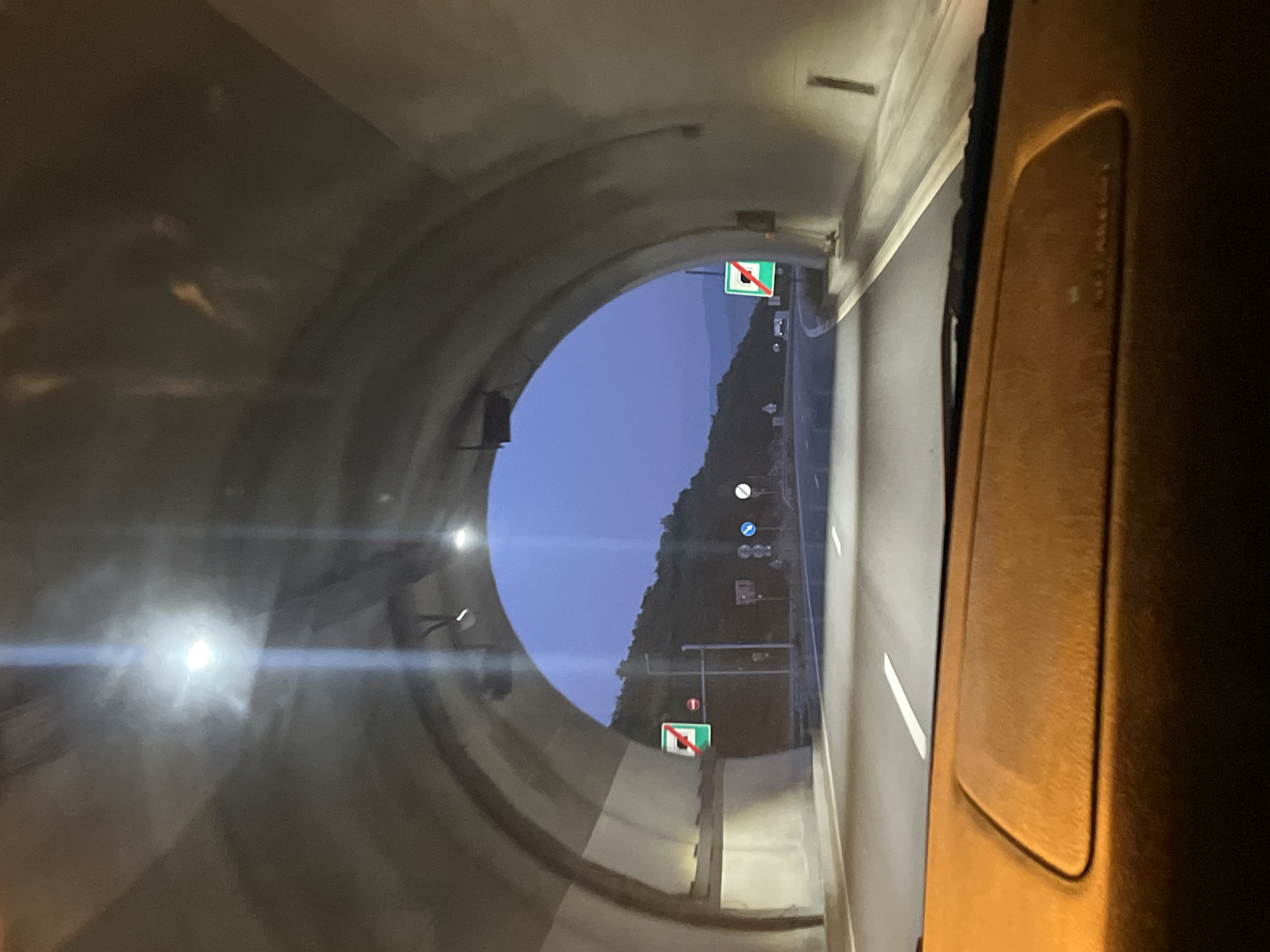 The image shows the interior of a vehicle driving into a tunnel. The tunnel has a concrete arched ceiling with lighting fixtures. On the left side of the tunnel, a green sign with a white square and a red diagonal line is visible, indicating the end of a restriction or a zone. Further down the road, there are other traffic signs and lights. The exterior of the tunnel opens to a hilly landscape under a twilight sky. The dashboard of the vehicle is visible in the foreground, suggesting the perspective is from the driver's seat. The road is paved and marked with white lines. The overall scene depicts a transition from daylight to the darkness of a tunnel.