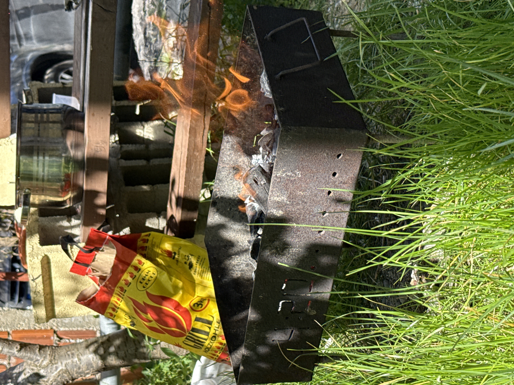 The image depicts an outdoor scene with a charcoal grill in use, situated in a grassy area. The grill is constructed from dark metal with visible signs of wear and tear, containing burning charcoal inside. Flames are actively rising from the grill's contents. Beside the grill, there is a partially torn yellow bag labeled "GRILL HOLZKOHLE" (grill charcoal in German). In the background, there are brick structures and a wooden fence. The presence of a car tire suggests the scene is set in a residential area or backyard. While it can be deduced that the grill is in operation, there is no immediate evidence to confirm if the location is the city of Bar, Montenegro. Additional information is required to confirm the location.