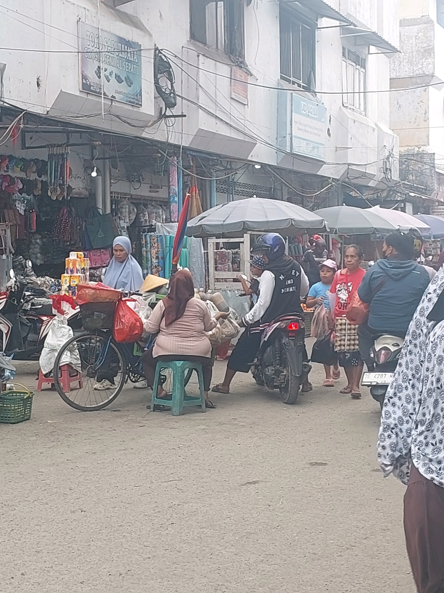 The image depicts a bustling street market scene in Ambon, Indonesia. The foreground features a street lined with market stalls selling various goods, including clothing, shoes, and what appear to be household items like toilet paper. Several people are visible, engaging in shopping and street activities.
In the center, a woman in a light blue hijab sits behind a bicycle laden with bags and merchandise, appearing to be a vendor. Beside her, another woman in a brown hijab is seated on a green stool. A motorcycle with two riders is moving through the scene, one of whom is wearing a helmet and a jacket with lettering. Further back, other individuals are seen browsing or walking.
The market appears to be situated in a built-up area, with buildings lining the street, some with awnings over the shopfronts. The overall atmosphere suggests a lively commercial district. The presence of motorcycles is notable, indicating common transportation in the area. The ground is paved and appears to be a street or walkway. The lighting suggests it is daytime, possibly overcast.