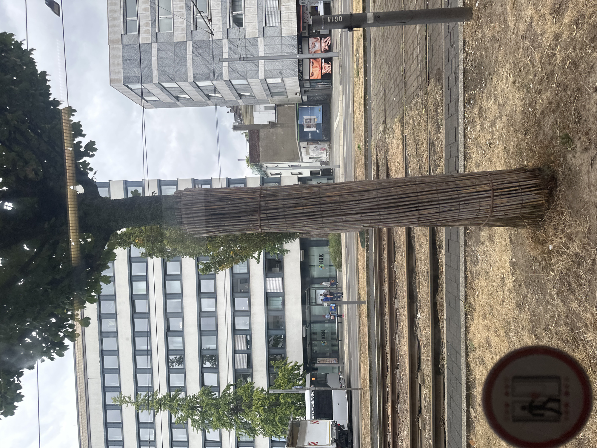 The image depicts a street scene in Krefeld, Germany, with a tram track in the foreground. A tree wrapped in bamboo stakes stands prominently in the middle of the frame. Behind the tree, a white bus is stopped at a bus stop beneath a modern building with multiple windows. The bus stop has a bench and signage. Further back, another building with a gray, patterned facade is visible, featuring shops at street level, including a nail salon with advertisements. The sky is overcast, and the overall environment suggests an urban setting on a cloudy day. A red circular sign with a pictogram of a person entering an elevator is visible in the lower left corner.
