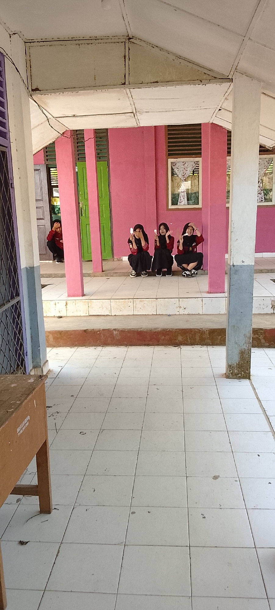 In Tanjung Setia, Indonesia, a group of four individuals, three females and one male, are visible in the outdoor corridor of a building. The three females are seated cross-legged on a tiled floor, appearing to be posing for a photograph. They are wearing dark hijabs and red tops with black bottoms, and each is holding a small white object, possibly a piece of ice cream or a treat. Their postures suggest they are interacting with each other and looking towards the camera. The male individual is partially visible in the background, crouching in front of a doorway, with his hand on his head, as if he is disengaging from the group or in a contemplative state. The environment features pink walls, green doors, and white tiled flooring. A wooden table or desk is present in the foreground on the left side of the image. The overall scene appears to be a casual gathering in front of a school or institutional building.