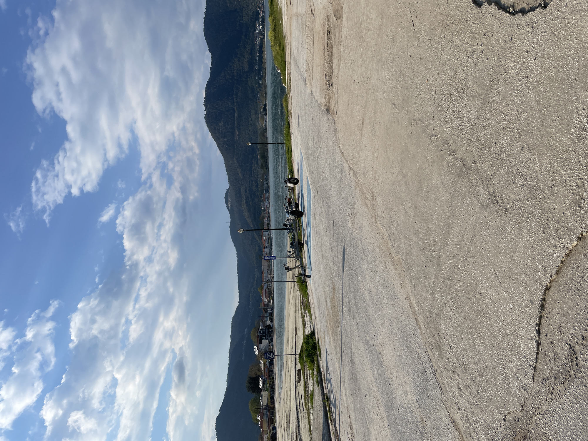 The scene is a street view in Skala Potamias, Greece, oriented in a rotated perspective. The background is comprised of mountains and a body of water, likely the sea. In the midground, street lamps line the waterfront pathway. Several scooters are parked near the waterfront. The foreground is primarily occupied by a concrete walkway or road. The sky is visible, featuring a blue hue with scattered clouds.