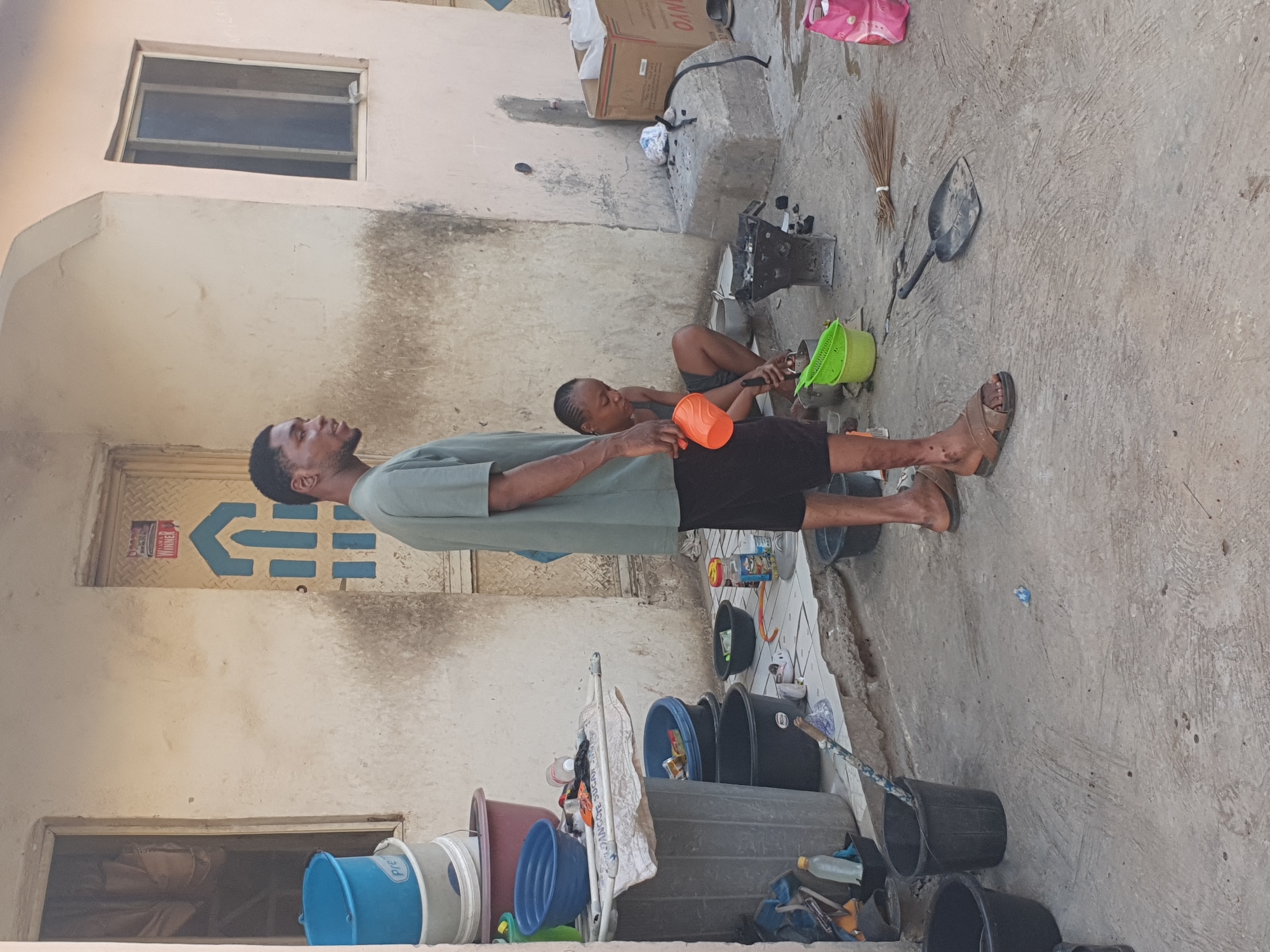 The image depicts an outdoor scene in what appears to be Lokoja, Nigeria. A man is standing with a woman sitting on the ground next to him. They both appear to be working, handling a green and an orange bucket. The area is cluttered, with various items like a cooking stove, a broom, a shovel, and numerous plastic buckets of different colors and sizes scattered around. The setting appears to be a backyard or courtyard, with weathered walls and a window visible. There are other items, including a box and what appear to be some cleaning supplies.