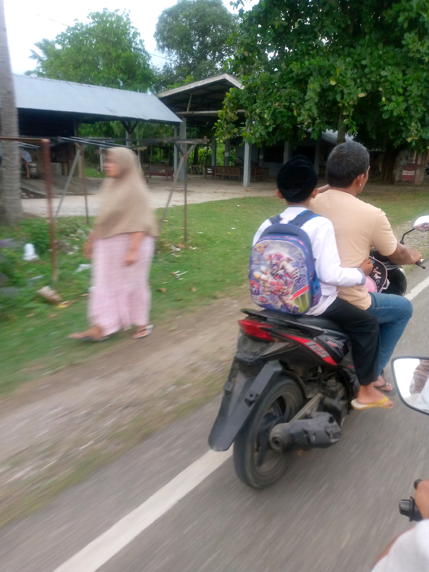 This image captures a scene in Glokaron, Indonesia, focusing on a motorcycle carrying two individuals. A person in the foreground, viewed from behind, is riding the motorcycle, wearing a light brown t-shirt and dark blue jeans. A younger individual, also viewed from behind, is seated in front of the rider, wearing a white long-sleeved shirt and black pants, with a colorful backpack featuring animated characters. The motorcycle is black and appears to be in motion on a paved road with a white line marking the lane. In the background, a woman in a light brown headscarf and a pink patterned skirt is walking on a grassy area next to a dirt path. Behind her, structures with corrugated metal roofs and wooden supports are visible, suggesting a rural or semi-urban setting with some vegetation. The overall atmosphere is outdoors, with trees and greenery present.