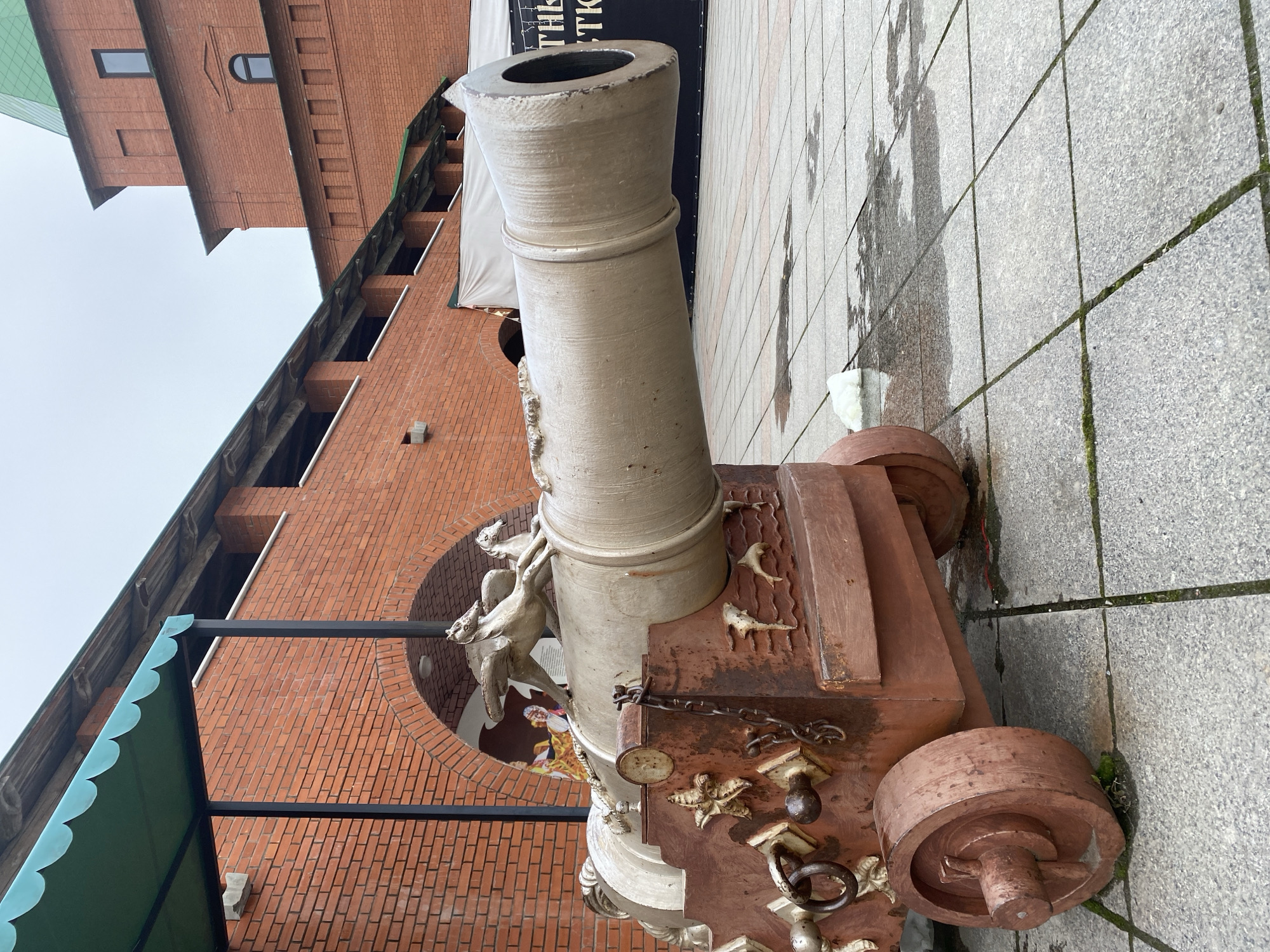 This media file captures a large, elaborately decorated cannon displayed outdoors on a wet, grey tiled pavement, with a multi-story red brick building serving as its backdrop.
The central subject is the cannon, featuring a light-colored barrel with distinct rings, suggesting it's made of stone or painted metal. Its carriage is a dark, reddish-brown material, possibly painted wood or metal, and is heavily adorned. Intricate sculptures of marine life, including starfish and stylized fish or sea creatures, are integrated into the design. A larger, figure resembling a mermaid or mythical creature supports the cannon's barrel at its base. Chains, rings, and other metallic embellishments are attached to the carriage, which rests on solid, reddish-brown wheels.
The setting includes a prominent red brick building with varied window shapes, including rectangular and arched designs. A green, scalloped awning extends from the building on the left. Behind the cannon, a semicircular archway in the brick wall reveals a colorful mural or image depicting a person in what appears to be historical or theatrical attire. A dark banner or sign is partially visible behind the cannon's barrel, displaying fragmented white text that includes "THIS" and Cyrillic characters such as "ТК".
The grey paved ground, showing moss in its grout lines, appears wet and reflects the overcast sky, suggesting recent rain or damp conditions. The sky is uniformly light grey, indicating an overcast day. No specific activity or event is taking place; the scene depicts a static architectural and sculptural display. The location is Yoshkar-Ola, Russia.