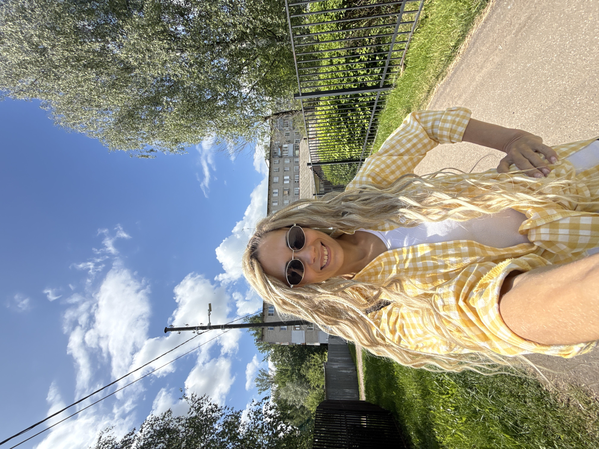 This media file presents a bright, outdoor selfie of a smiling woman in what appears to be a residential or park setting in Rusanovo, Russia. The woman, with long, wavy blonde hair and round dark sunglasses, is looking directly at the camera with a joyful expression. She is wearing a yellow and white gingham plaid button-up shirt over a white top.
The scene is illuminated by bright daylight, under a clear blue sky dotted with white, fluffy clouds, indicating sunny and pleasant weather. In the background, a paved path runs alongside a stretch of green grass and lush trees. A black metal fence is visible, separating different areas of greenery. Further back, a multi-story, grey residential building with numerous windows suggests an urban or suburban environment. A utility pole with several wires stands out against the sky, adding to the infrastructural elements of the setting. The overall impression is one of a cheerful, casual moment captured outdoors. There is no visible text in the image.