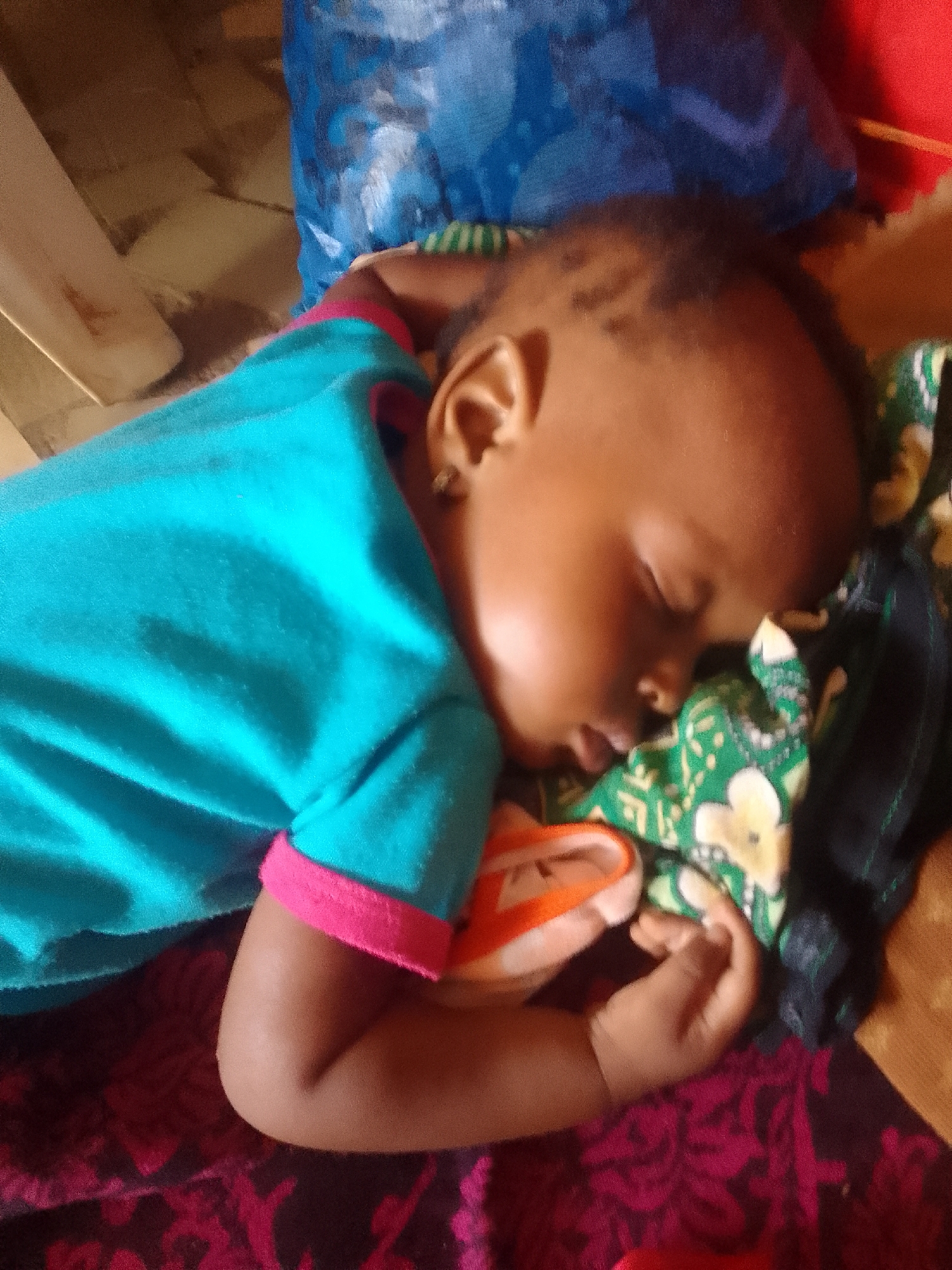 A young child is sleeping soundly on their side, facing the viewer. They are wearing a turquoise t-shirt with pink trim. The child's right arm is tucked under their head, and their left hand is resting on their chest. The child's eyes are closed, and their mouth is slightly open, suggesting deep sleep.
The child is lying on a patterned fabric, which appears to be a blanket or bedding. To the child's left, there's another patterned fabric, predominantly green with white floral designs and some black elements. Behind the child, a large blue object, possibly a bag or cloth, is visible. The background is out of focus, but appears to be an indoor setting with some wooden elements and a tiled floor. The lighting suggests it might be daytime. The overall impression is one of peaceful rest. The location is Sabon Tasha, Nigeria.