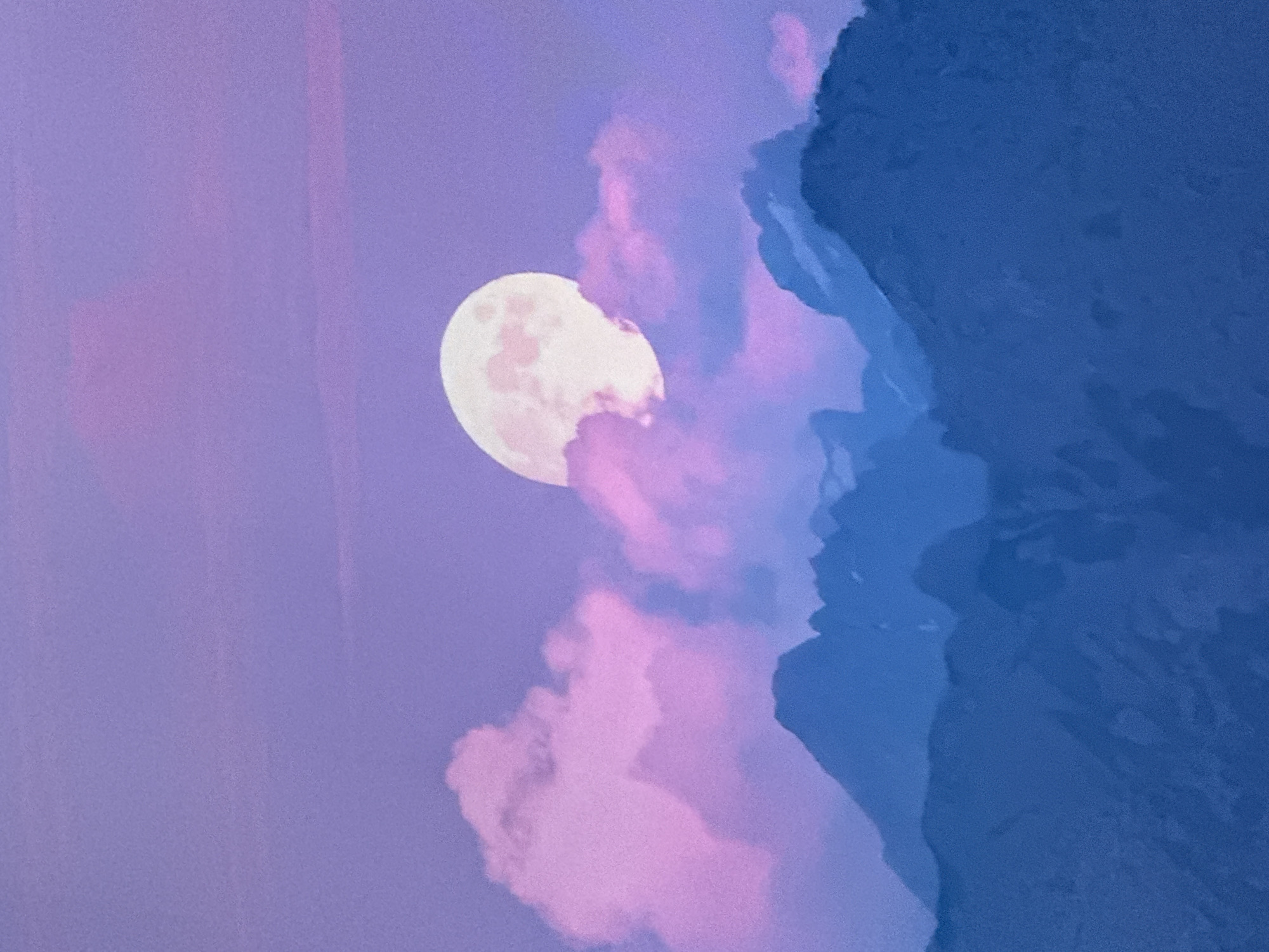 The image shows a full moon rising behind a range of silhouetted mountains. The sky is a gradient of deep purple to lighter lavender, with wispy pink clouds scattered around the moon. The mountains are dark blue and appear rugged and steep, with hints of snow or light-colored rock visible on some of the peaks. The overall mood of the image is serene and ethereal, with the soft colors and the glow of the moon creating a dreamlike atmosphere. The image does not depict any elements related to the city of Simferopol, Ukraine.