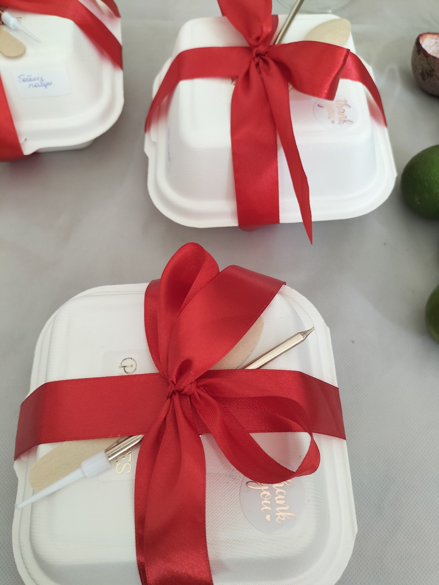 This image features a close-up view of three white, compostable food containers, each adorned with a bright red satin ribbon tied in a bow. Two of the containers are clearly visible, with the third partially obscured in the background. Each container also holds a wooden utensil and a small metallic-colored utensil with a white tip.
On the top left container, a small white label is affixed, containing handwritten text that appears to be "Főztük nagy". On the middle container, a circular sticker with the text "Thank you" in a cursive font is visible. A similar "Thank you" sticker is present on the bottom container, though it is partially covered by the ribbon.
The setting appears to be an indoor event or gathering, likely a catered meal or a gift presentation. The neutral background suggests a table setting. The presence of limes and what looks like the rind of a passion fruit in the background might hint at the type of food or beverages being served. The lighting is soft and even, suggesting an indoor environment, possibly during the day. There are no people visible in the frame, focusing the attention solely on the beautifully presented food containers. The overall impression is one of thoughtful preparation and a welcoming gesture.