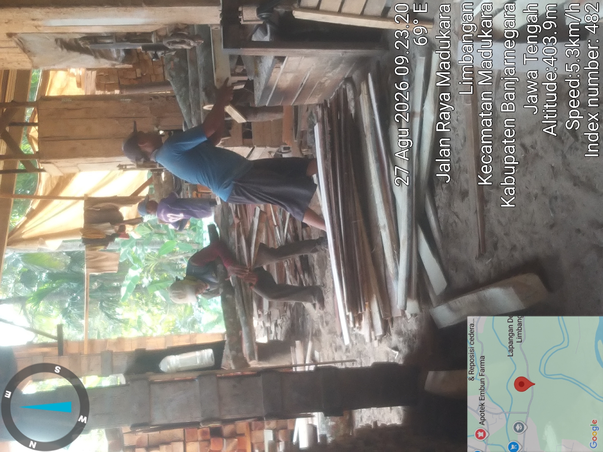The image shows an outdoor industrial scene with several people working with wood. In the foreground, a person in a blue shirt and dark skirt is operating a woodworking machine, feeding logs into it. Another person in a blue shirt and dark pants is standing behind a pile of lumber, and a third person wearing a light-colored cap and vest is carrying a stack of wooden planks. The environment appears to be a workshop or lumberyard, with piles of cut wood and machinery visible. The background shows trees and foliage, suggesting a rural or semi-rural setting. A compass rose graphic is superimposed on the upper left corner of the image, indicating North, East, South, and West. Text overlays on the image provide location and time details, including "27 Agu 2026 09.23.20 69° E", "Jalan Raya Madukara Limbangan Kecamatan Madukara Kabupaten Banjarnegara Jawa Tengah Altitude:403.9m Speed:5.3km/h Index number: 482", and "Apotek Embun Farma & Reposisi cedera...". A Google Maps inset in the lower left corner shows a map with a red marker indicating a location within Limbangan.