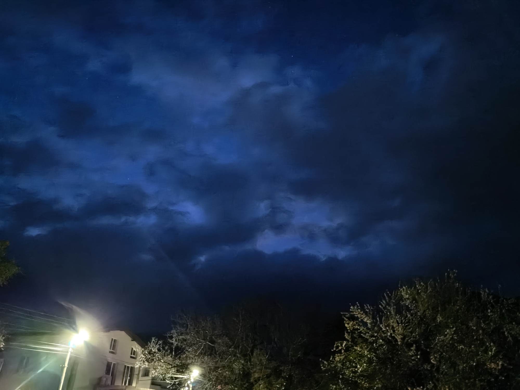 The image shows a night scene in an urban environment. The sky is dominated by dark, blue-grey clouds, with small points of light that may be stars. The lower portion of the image depicts the silhouette of trees and a building lit by streetlights. The building appears to be residential, with visible windows. The overall environment suggests a quiet, residential area at night. The location is in the city of Unknown, Unknown.