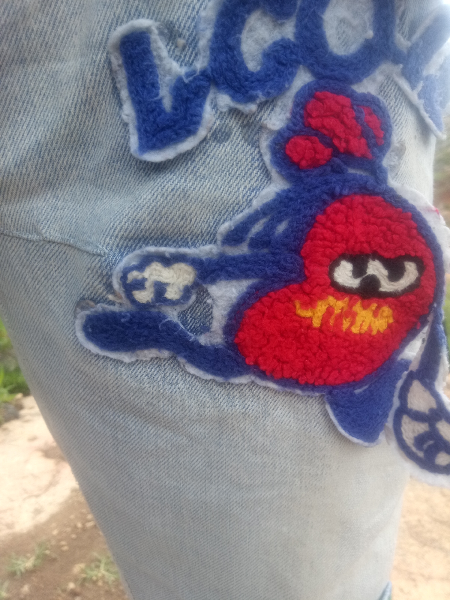 The media file displays a close-up, vertical shot of light blue denim fabric adorned with a colorful patch. The patch features a cartoonish character with a red, heart-shaped body outlined in blue. The character has two white eyes with black pupils and a yellow mouth. Its limbs are also blue. Above the character, partially visible, are blue embroidered letters with white outlines, suggesting it's part of a larger design, possibly a name or logo. The denim fabric appears textured with diagonal twill lines. The background is blurred, showing earthy tones of ground and hints of green vegetation, suggesting an outdoor setting. The lighting suggests it is daytime. The image focuses on the detail and texture of the patch and the denim.