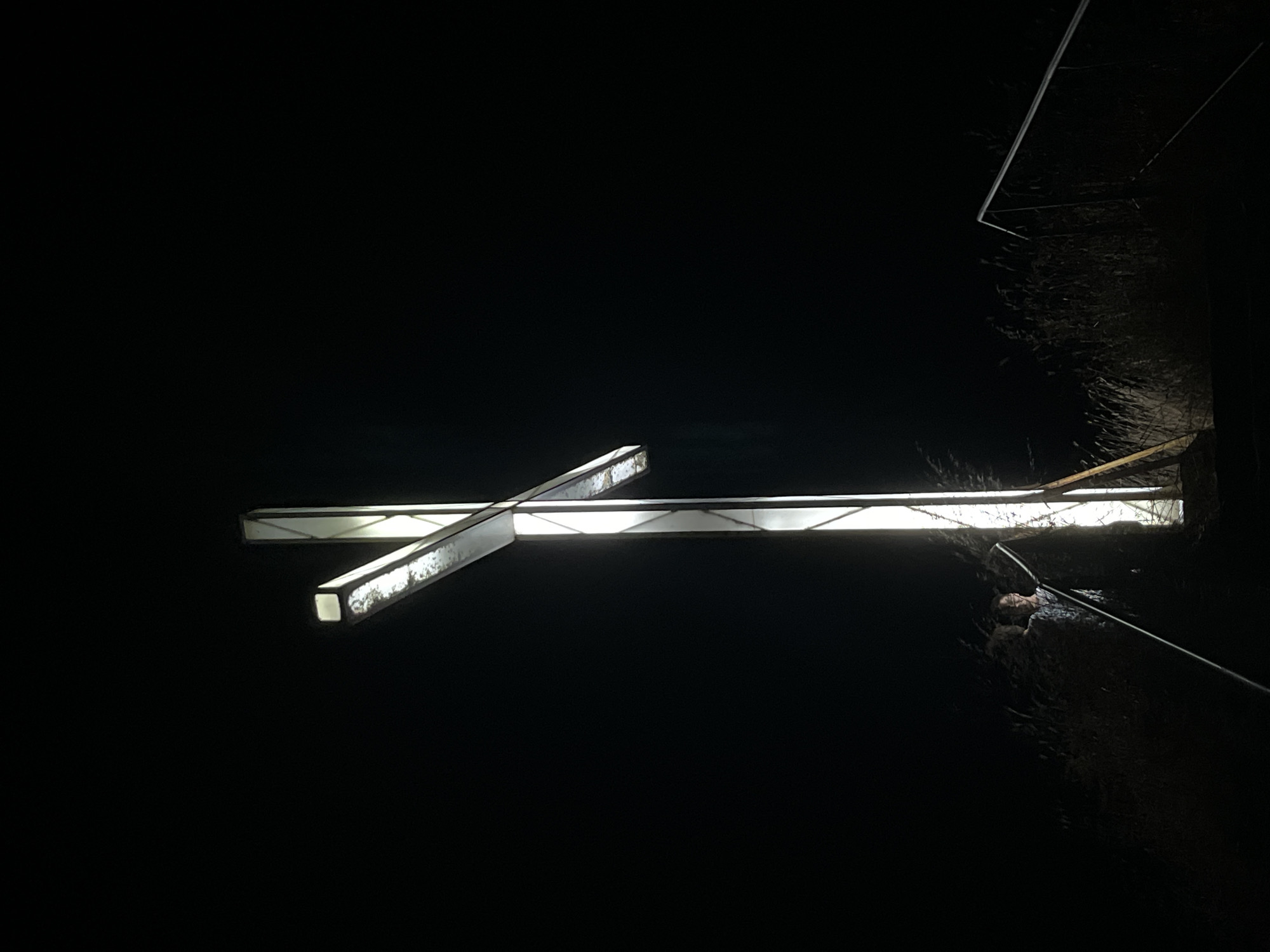 A person stands on stairs in front of a large, illuminated cross at night. The cross is constructed of white, translucent material and appears to be made of metal tubing with panels. It is lit from within, casting a bright glow against the dark night sky. The person, who is wearing a patterned shirt, is looking towards the cross and appears to be standing on a set of steps that lead up to the cross. There are railings on either side of the stairs. The area around the cross and stairs is overgrown with dry grass and reeds, suggesting a rural or undeveloped location. The image was taken in Lozenets, Bulgaria.