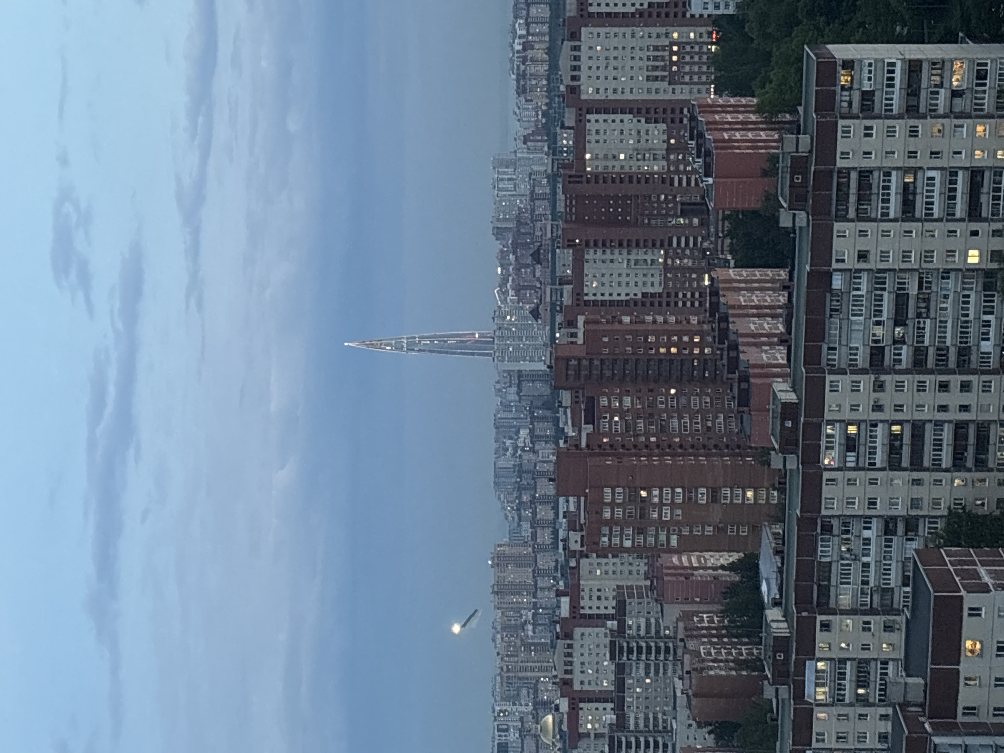 The image displays a panoramic city skyline at dusk or dawn, initially rotated 90 degrees clockwise. When correctly oriented, a light blue-grey sky with subtle cloud patterns fills the upper half.
The focal point is a very tall, slender skyscraper with a pointed top, positioned prominently in the mid-distance. Its internal lights are visible, highlighting its structure, strongly suggesting it is the Lakhta Center in St. Petersburg, Russia.
The foreground and midground are densely packed with numerous multi-story buildings, appearing to be residential or mixed-use structures. These buildings exhibit repetitive architectural patterns, typical of Brutalist or Soviet-era design, with facades in shades of grey, brown, and white. Many windows are illuminated, indicating occupancy and activity within the city.
In the mid-left portion of the sky, an elongated object with a distinct bright light on one end is visible, appearing to be an airplane or helicopter in flight. Patches of dark green foliage are interspersed among the buildings. The scene conveys the vastness of an urban environment during twilight. No specific individuals or events are captured, and no text is visible.
