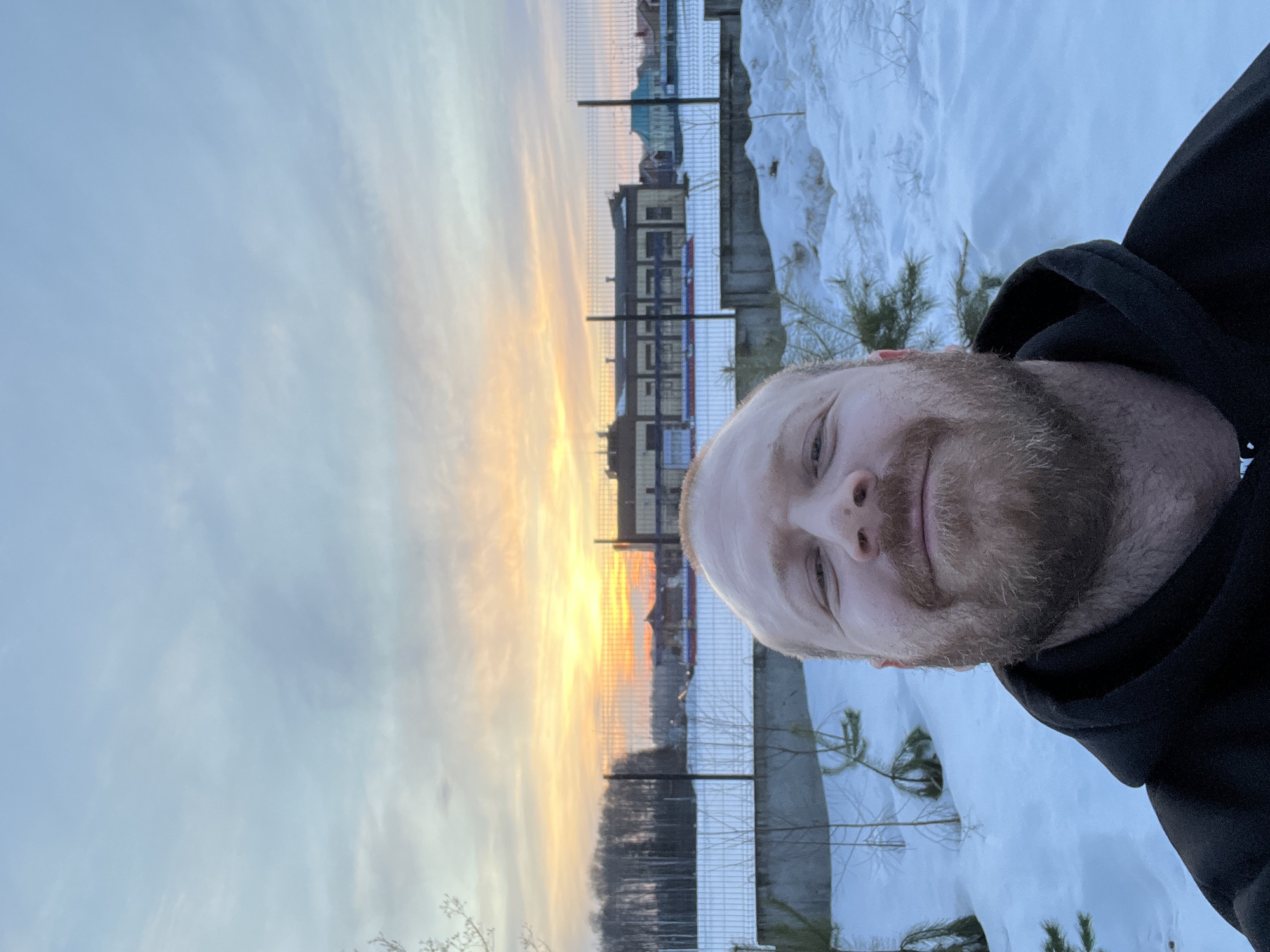 An adult male subject is depicted in a close-up selfie, occupying the lower half of the frame. He has fair skin, a reddish-brown beard, and is wearing a dark hooded sweatshirt. He smiles faintly while looking directly into the camera. The background reveals an outdoor, winter scene in Noviy, Russia, during either sunrise or sunset, characterized by an orange and yellow glow on the horizon transitioning into a pale blue and grey sky. The ground is covered in snow. A chain-link fence extends across the mid-ground. Beyond the fence, a large, multi-story industrial or commercial building with numerous windows is visible. To the left, a dense line of deciduous trees borders the horizon, while to the right, several residential structures with colored roofs are present in the distance. Small evergreen saplings are sparsely scattered in the snow in the foreground. A concrete barrier runs horizontally behind the foreground snow.
