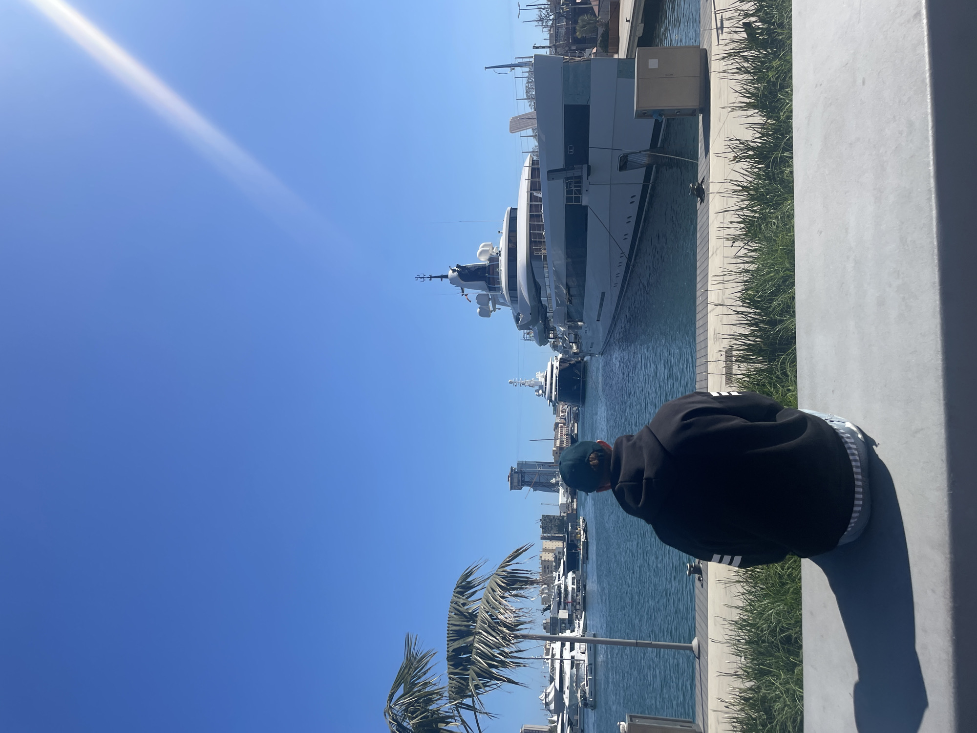 A partially visible individual in a black hoodie and green baseball cap sits on a gray concrete ledge, overlooking a harbor in Barcelona, Spain. In the background, several yachts and boats are docked, including a particularly large white multi-story yacht. Palm trees sway in the foreground. The sky is clear blue, with a bright streak, possibly sunlight reflecting off an object. Buildings are visible in the distance. The water in the harbor appears calm.