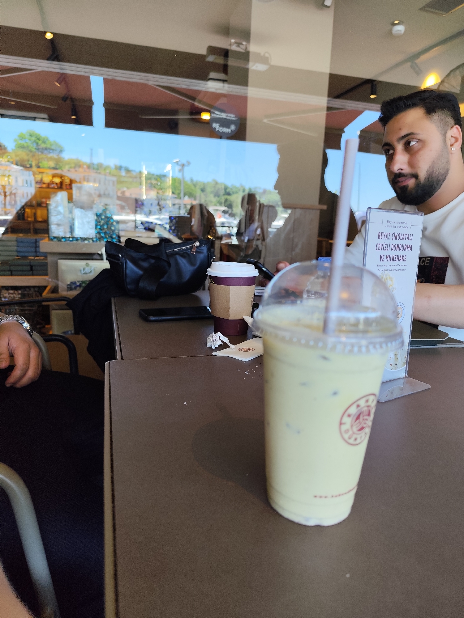 The image captures a scene in what appears to be a café in Sarıyer, Turkey. In the foreground, a plastic cup of a beverage, likely a milkshake, sits on a brown table. A straw is inserted into the cup. Beside it, there is a coffee cup, and a cell phone. In the background, a black handbag is visible, along with retail displays and packaging. Through the window of the café, a daytime cityscape can be seen with buildings, trees, and what may be water.
To the right, a man is partially visible in the frame. He is wearing a white shirt and has dark hair and a beard. He appears to be looking towards the right of the frame. Next to him, there is a card stand featuring text in Turkish about "BEYAZ ÇİKOLATALI CEVİZLİ DONDURMA VE MILKSHAKE" (White Chocolate Walnut Ice Cream and Milkshake). Another person’s hand with a watch is visible in the left foreground. The overall lighting of the scene suggests it is daytime, possibly mid-day.