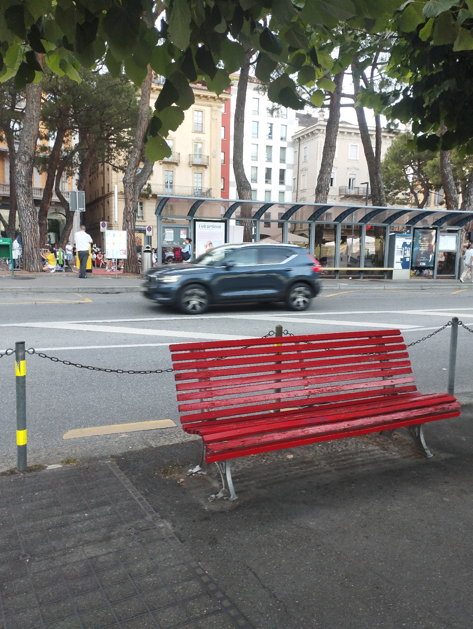 The image captures a street scene in Lugano, Switzerland, likely near a bus stop. In the foreground, a bright red wooden bench with metal legs sits on a paved area. A metal chain fence with posts topped with yellow and grey markings separates the bench area from the road. The road itself has white lane markings and a pedestrian crossing.
In the midground, a dark blue car is in motion, blurred due to its speed. Behind the road, a modern bus stop shelter with a curved glass roof and metal frame is visible. Several people are present at the bus stop, some standing, others appearing to be seated. Behind the bus stop, multi-story buildings with architectural details like balconies and numerous windows line the street. Trees with lush green foliage are scattered throughout the scene, some obscuring parts of the buildings. The overall lighting suggests it is daytime, possibly late afternoon or early evening.