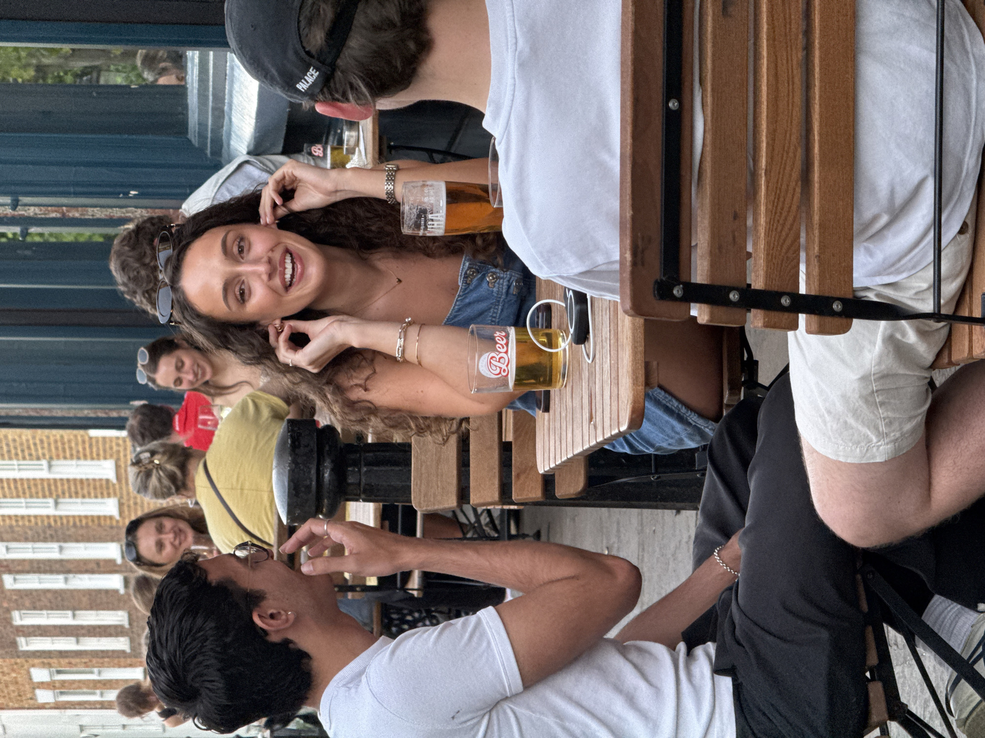 A group of people are sitting at a table outside, presumably at a pub or restaurant. They are enjoying drinks and conversation. One woman with curly brown hair is smiling at the camera, holding her ear. Several other people are visible in the background, including a man wearing a black cap with "PALACE" written on it. The background shows buildings that suggest this could be in London, United Kingdom.