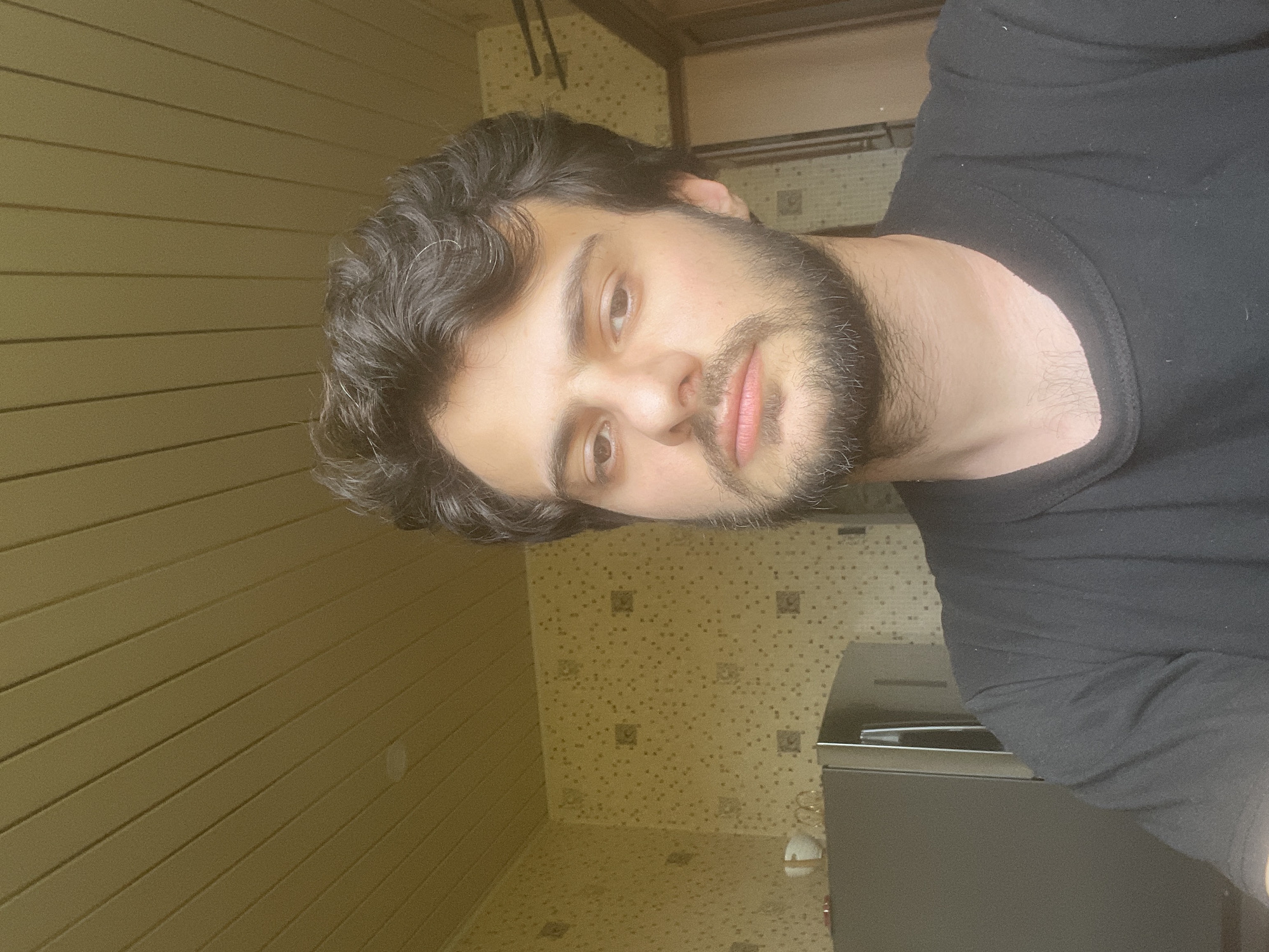 A medium shot captures a young adult male with dark, wavy hair and a beard, wearing a black t-shirt. He is indoors, with a textured, yellow ceiling and patterned wallpaper visible in the background. To his left, a stainless steel refrigerator is present. The lighting suggests a daytime setting, with shadows indicating light sources from the upper left. The scene appears to be a residential kitchen or living area. The specific location in Tbilisi, Georgia, is not discernible from the visual information provided.