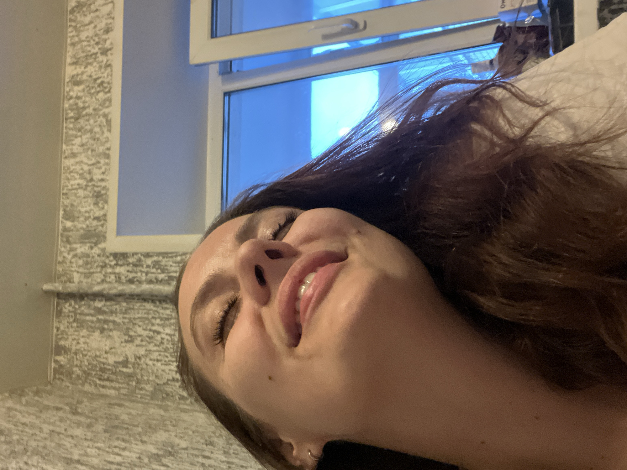 The media file presents an intimately framed, angled portrait of a young woman, appearing to be lying down and looking upwards. Her head is tilted to the left, with her dark brown hair flowing around her. Her eyes are closed, and her lips are parted in a gentle smile, revealing her upper teeth, conveying a sense of serenity, contentment, or joy. A small mole is visible on her right cheek.
The background primarily features a large, white-framed window, through which a bright, diffused blue light is visible, indicative of daytime. To the left of the window, a wall section exhibits a textured, mottled grey pattern, possibly decorative wallpaper or plaster, bordering a plain light grey wall. A hint of a light-colored object, likely a pillow or bedding, is visible near her head on the right side of the frame.
The image captures a moment of relaxation or rest in an indoor setting, presumably a home or hotel room. There is no visible text. Given the context, this scene is situated in Chaikovsky, Russia.