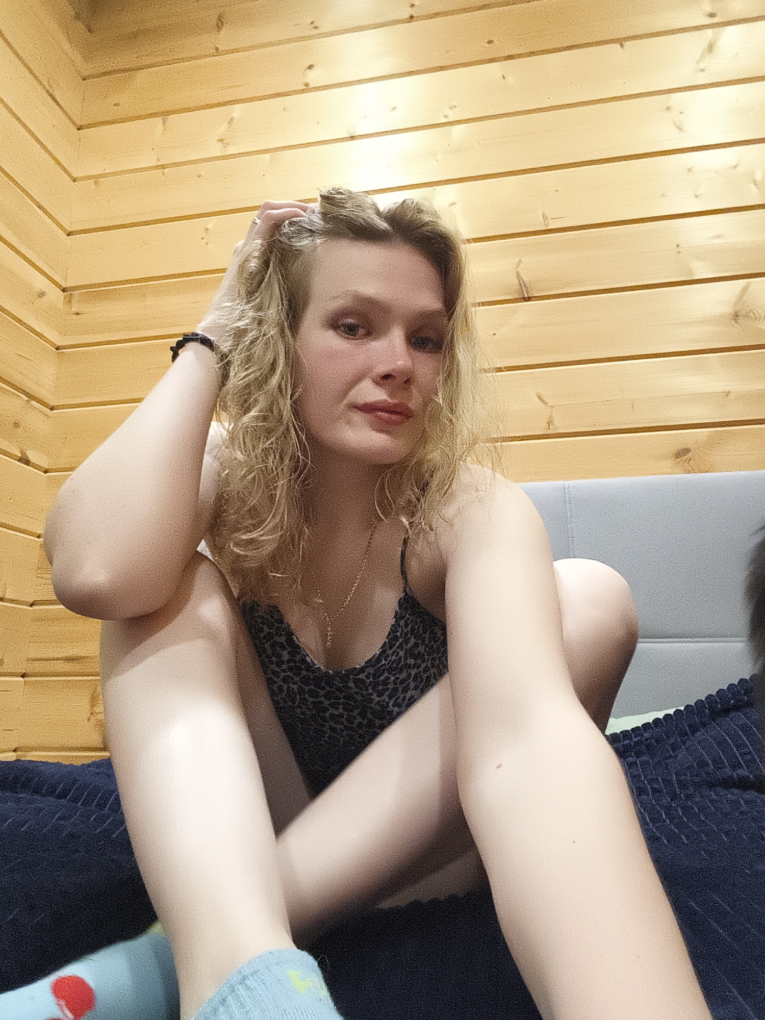 A woman with curly blonde hair is sitting on a bed with her legs crossed. She is wearing a spaghetti strap top with a black and gray cheetah print pattern. Her right hand is touching her hair, and she has a black bracelet on her wrist. She is wearing light blue socks. The bed has a gray headboard and a dark blue, textured blanket. The walls are covered in light brown wooden planks. The photograph appears to be taken indoors in Русский Вожой, Russia.