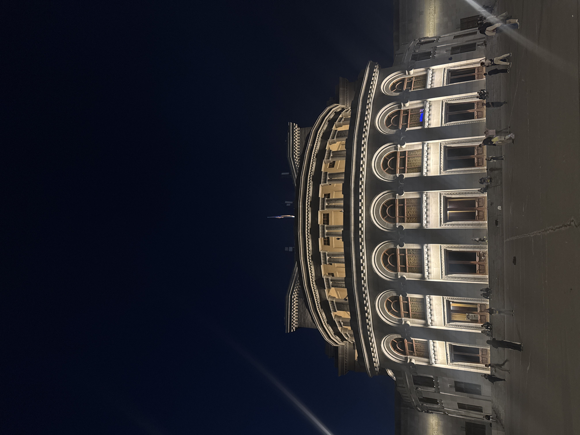 The image shows the National Gallery of Armenia in Yerevan at night. The building is a large, neoclassical structure with a curved facade. Arched windows and columns are visible. People are walking in the plaza in front of the building. The sky is dark.