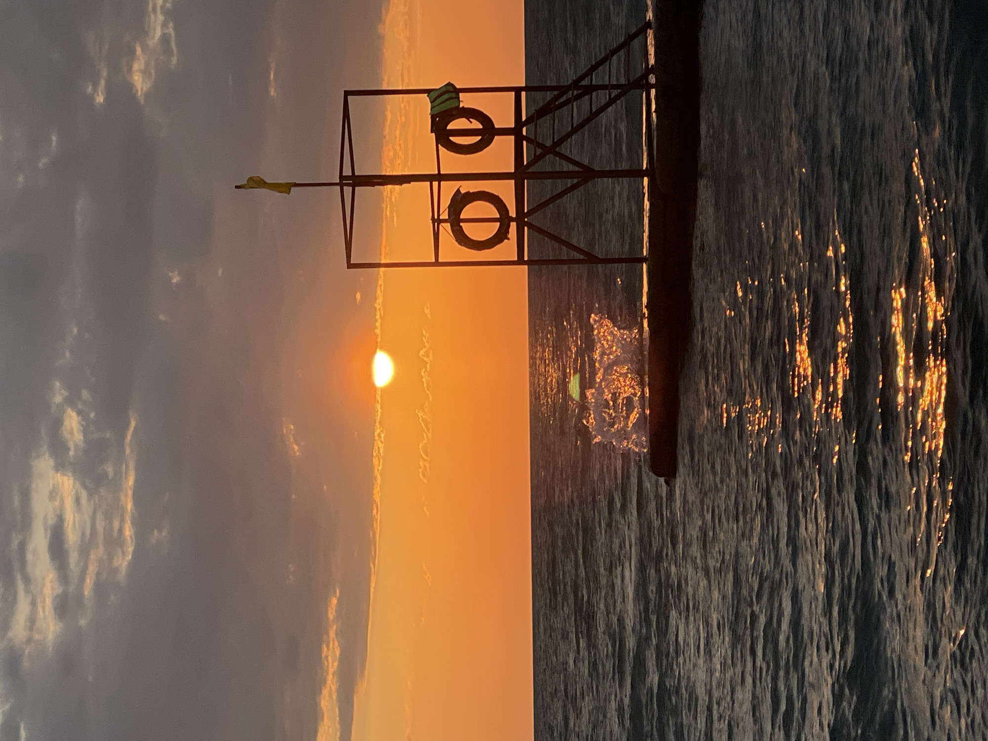 A sunset scene is captured over the water, likely in or near Bogushevka, Russia, judging by the provided context. The sun, a bright orb, is partially obscured by clouds, casting a warm orange and yellow glow across the sky and reflecting on the water's surface. A dark silhouette of a lifeguard tower stands on a pier, equipped with two life preservers and a yellow flag at the top. The water is choppy, with a splash of white visible near the pier, illuminated by the setting sun. The overall atmosphere is serene and highlights the natural beauty of the evening.