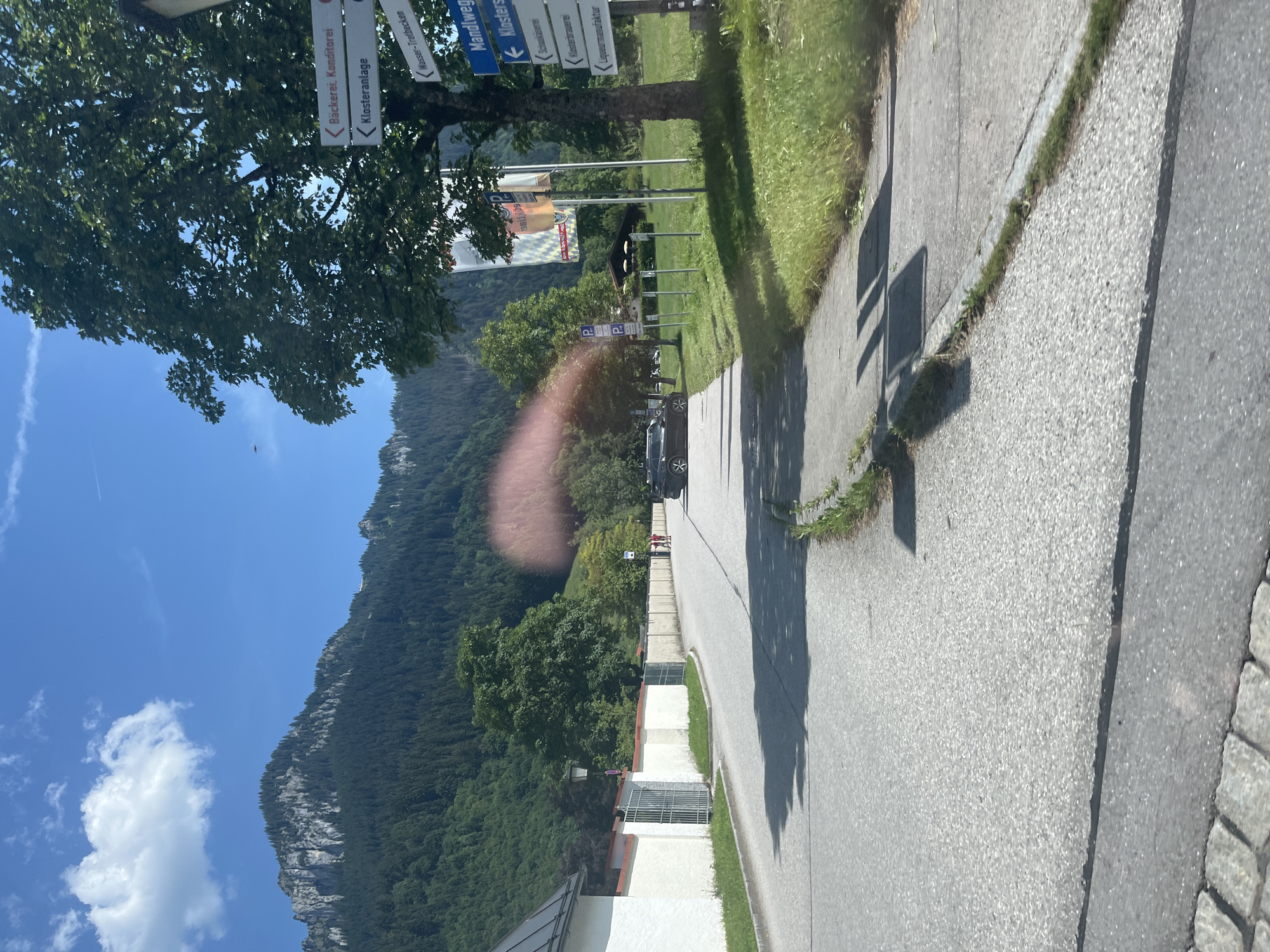 The image captures a scene in Oberau, Germany, featuring a road leading towards a mountainous backdrop. A white building with a dark roof is visible on the left, bordered by a white wall with a red trim and metal gates. A paved sidewalk runs alongside the road. On the right side, a large tree stands prominently, with several directional signs attached to its trunk. These signs indicate directions to "Bäckerei, Konditorei" (Bakery, Confectionery), "Klosteranlage" (Monastery Complex), "Mandlweg" (Mandl Path), "Schlossbrauerei" (Castle Brewery), and "Likörmanufaktur" (Liqueur Manufactory). Further down the road, two individuals are walking, and a dark-colored car is parked. The background consists of lush green trees covering the lower slopes of a rocky mountain range under a clear blue sky with scattered clouds. Parking signs are also visible on the right side of the road.