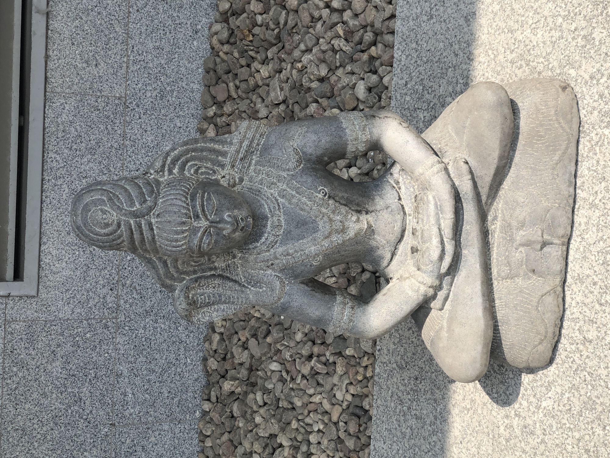 This is a photograph of a stone statue of a seated deity, likely Shiva, in a meditation pose. The statue is made of dark gray stone and depicts the deity with intricate carvings of jewelry and hair. A cobra is coiled around the deity's neck. The statue is placed on a light gray, speckled platform, in front of a wall made of similar speckled stone. Behind the statue, a bed of small, rounded river stones is visible. A metal vent or grate is present at the top of the wall. The lighting suggests it is daytime, with a shadow cast on the platform to the left of the statue. The image is taken from a frontal, slightly elevated perspective. The location of this statue in Warsaw, Poland, is the Temple of Polish Sadhana.