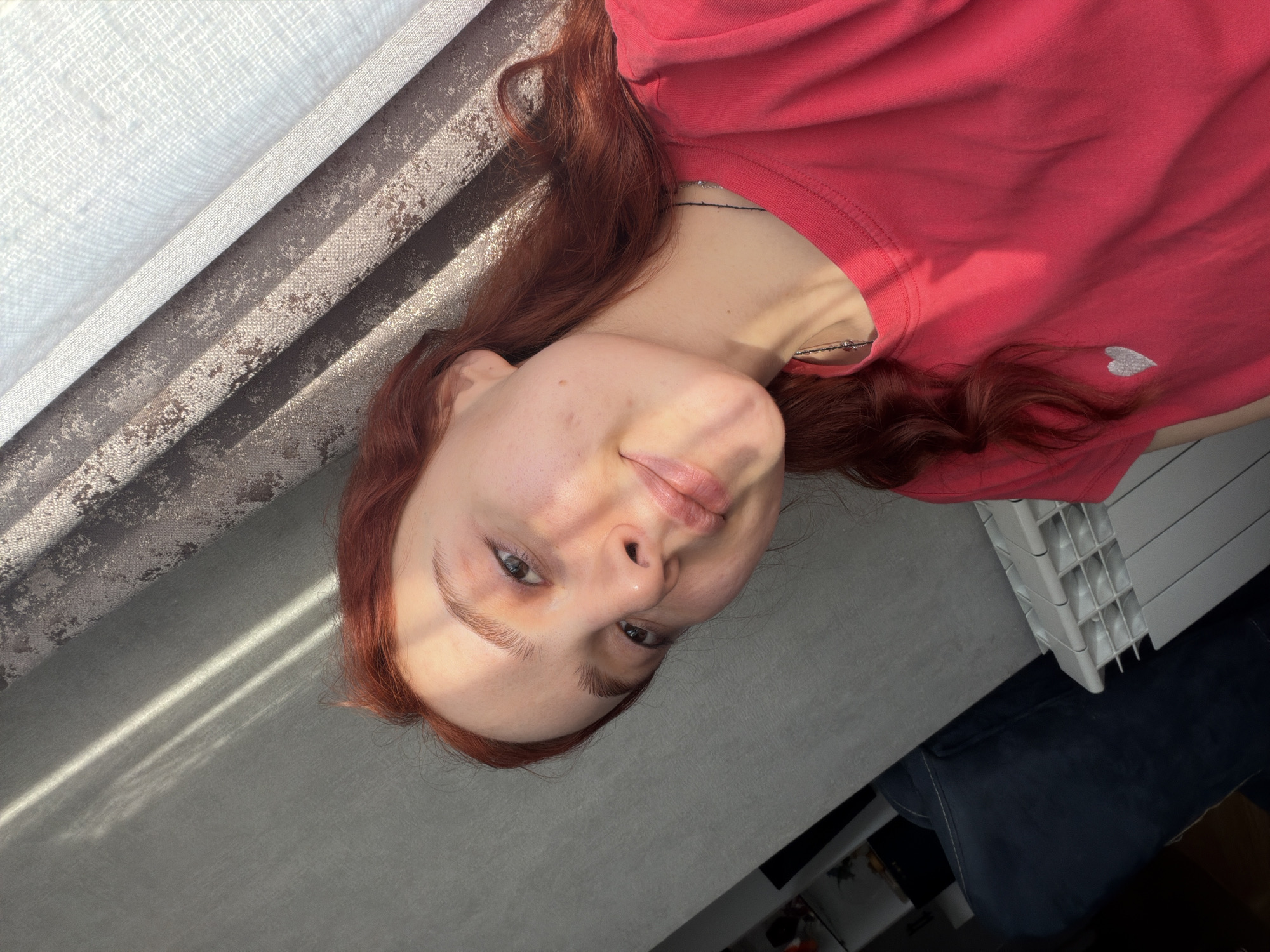 A young woman with auburn hair is lying down. She is wearing a coral-colored t-shirt with a small white heart on the lower right. The woman has fair skin, light eyes, and prominent eyebrows. The background includes a textured curtain or fabric in tones of white, gray, and brown. There is a gray surface with strong directional light. A white radiator and a dark fabric (possibly a blanket or couch) are also visible. The setting appears to be indoors, possibly in Tbilisi, Georgia, based on the overall aesthetic and architectural styles often found in the region.