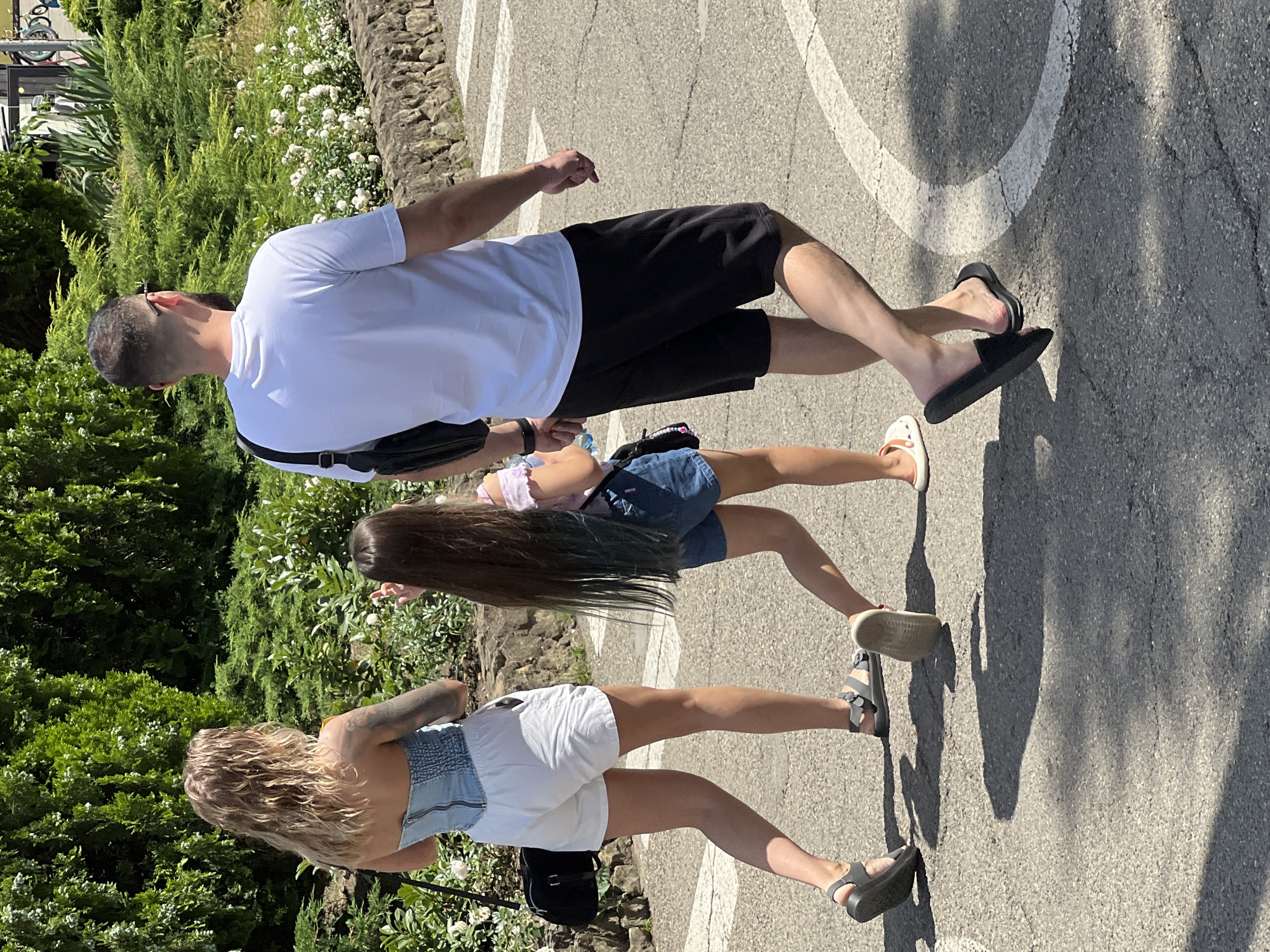 In Krasnodar, Russia, a man, woman, and child walk on a sunlit paved path lined with lush green bushes and flowering plants. The woman, with blonde, wavy hair and tattoos on her arm, wears a denim corset-style top and white shorts, carrying a black bag. The child, with long brown hair with green tips, is dressed in a light pink top and denim shorts. The man, in a white t-shirt and black shorts, carries a black bag on his shoulder. They are walking away from the camera, casting shadows on the ground.