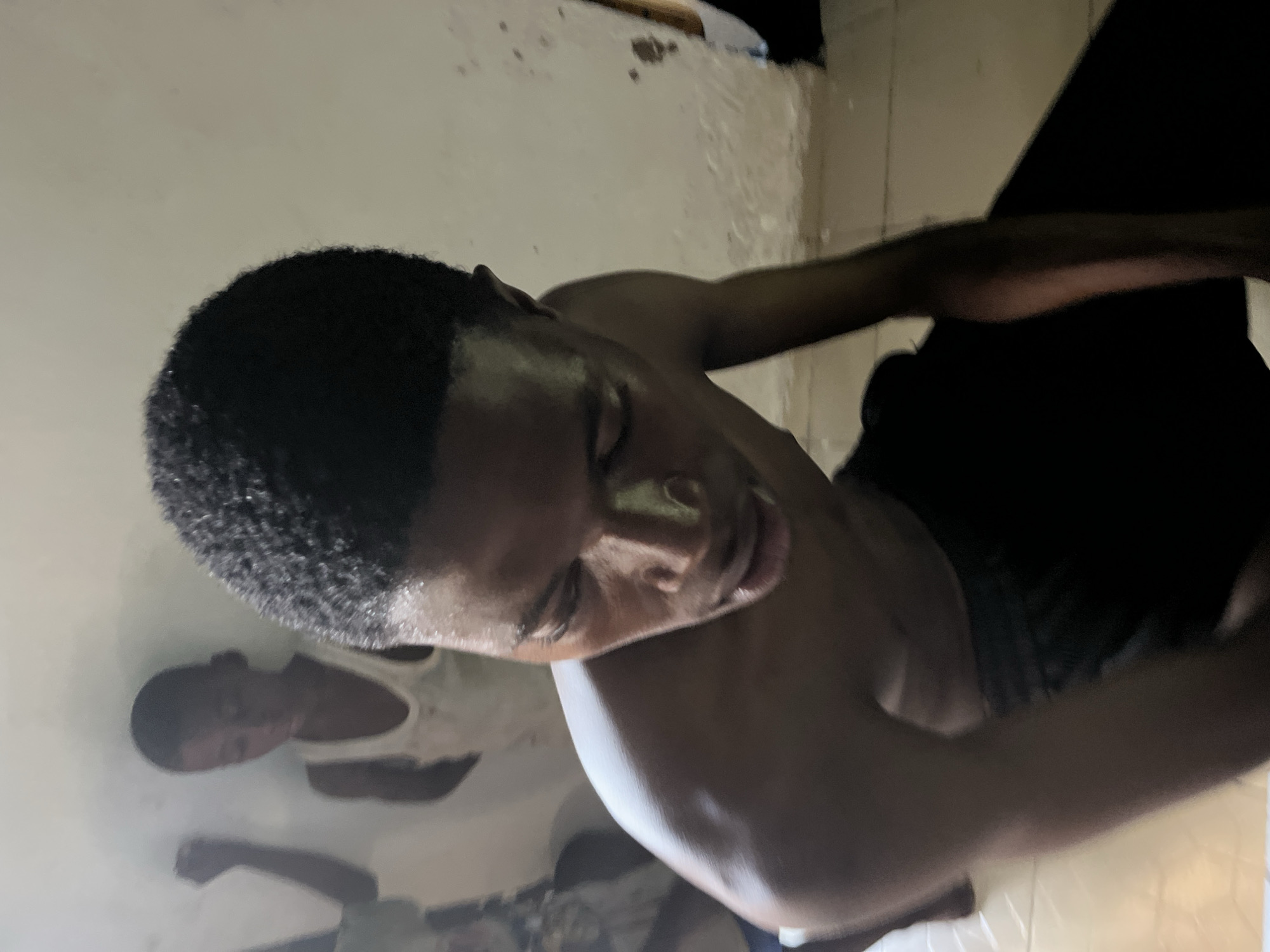 The media file shows a close-up, rotated view of a young Black man lying down. His eyes are closed, and his head is tilted back, revealing his neck and chest. His hair is short and textured. His right arm is bent and resting on his chest, while his left arm is extended outwards. Another person, also appearing to be a young Black male, is visible in the background, lying down and wearing a white tank top. The setting appears to be an indoor space with plain, light-colored walls and a tiled floor. The lighting is dim, creating shadows and highlighting the texture of the subjects' skin and hair. The overall mood appears to be one of rest or possibly exhaustion. There is no visible text, and the time of day and weather are not discernible.