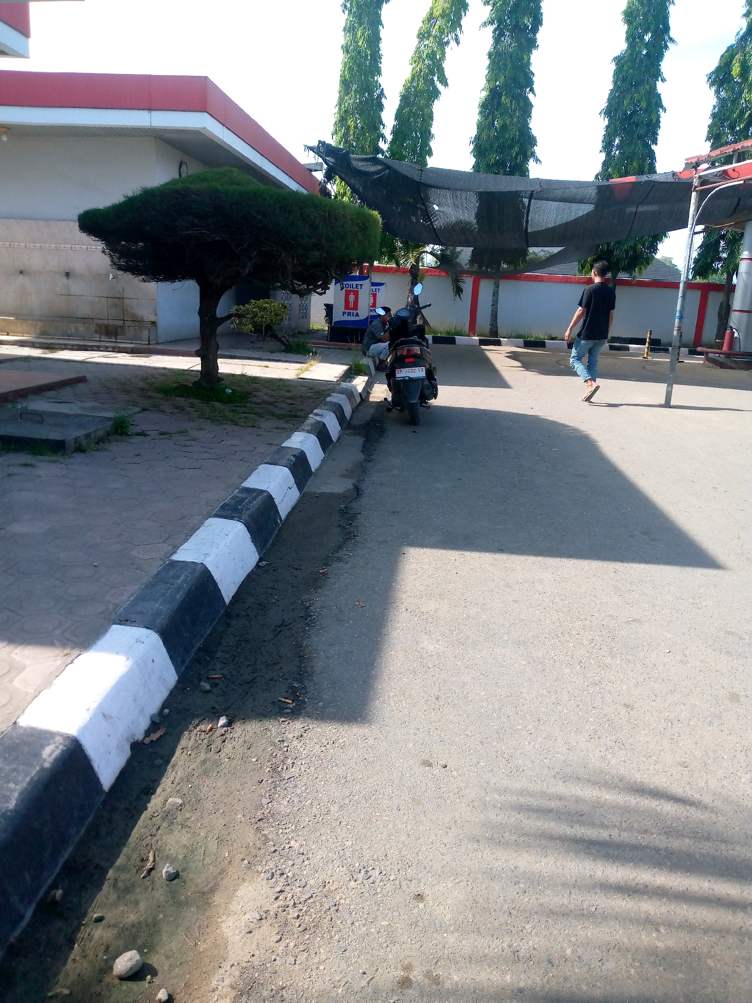 In Keudebayu, Indonesia, a black motorcycle is parked on a paved road beside a curb painted with alternating black and white stripes. A person in a black t-shirt and blue jeans walks away from the motorcycle towards the right of the frame, casting a long shadow. Behind the motorcycle, a sign indicates "TOILET PRIA" (Men's Toilet). To the left, a building with a red trim and white facade is visible, with a uniquely shaped trimmed tree in front of it. Tall trees with dense foliage line the background, some partially obscured by a dark mesh screen. The ground is a mix of pavement and dirt, with scattered small rocks and debris.