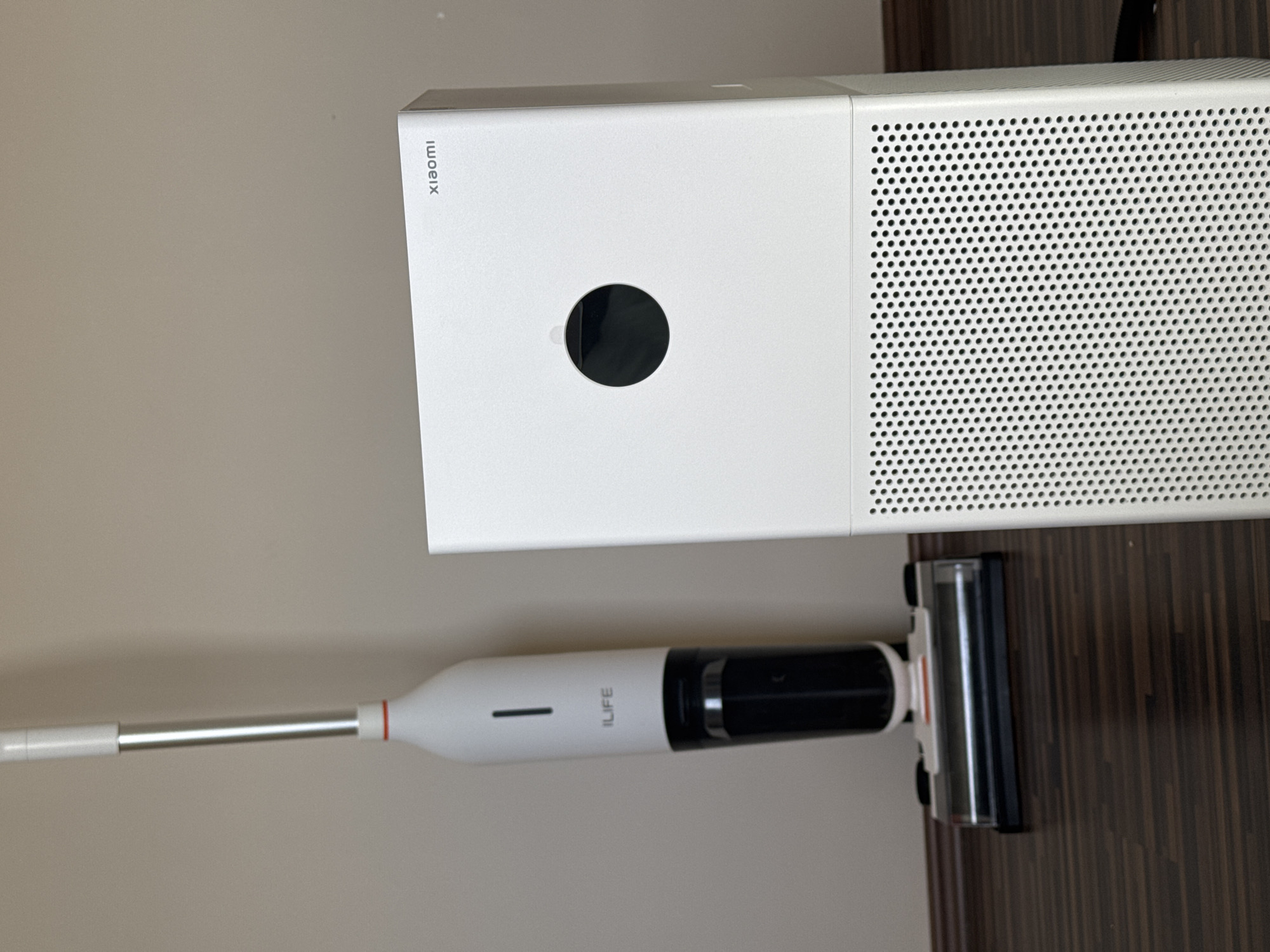 The image shows a white Xiaomi air purifier and an iLife vacuum cleaner. The air purifier is rectangular with a perforated surface on one side and a circular opening on the front. The vacuum cleaner has a white handle, a silver extension, and a black body with the "iLife" brand name printed on it. The floor appears to be dark brown. The location is not determinable based on the image.