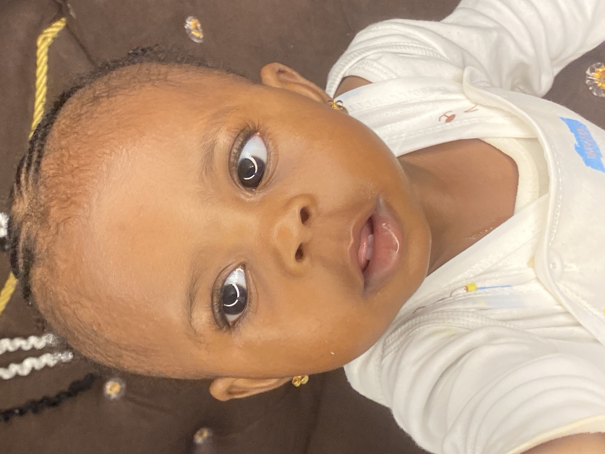A close-up, head and shoulders studio portrait depicts a young Black child, appearing to be less than two years old, looking slightly to the right of the camera. The child has dark skin, dark eyes, and dark hair styled in thin braids, with some loose wisps around the forehead. The child is wearing a white long-sleeved shirt with ribbed detailing. A small, gold, bow-shaped earring is visible on the child's right earlobe. The background is a dark brown, textured surface, possibly upholstery, with some blurred, sparkling elements that might be decorative. The lighting is soft and directed, highlighting the child's facial features and skin. The image is sharp and focused on the child's face, with the background softly blurred. The overall impression is that of a posed portrait taken in a controlled environment, likely a home or studio setting. There are no identifiable landmarks or context to suggest a specific location within Abakaliki, Nigeria.