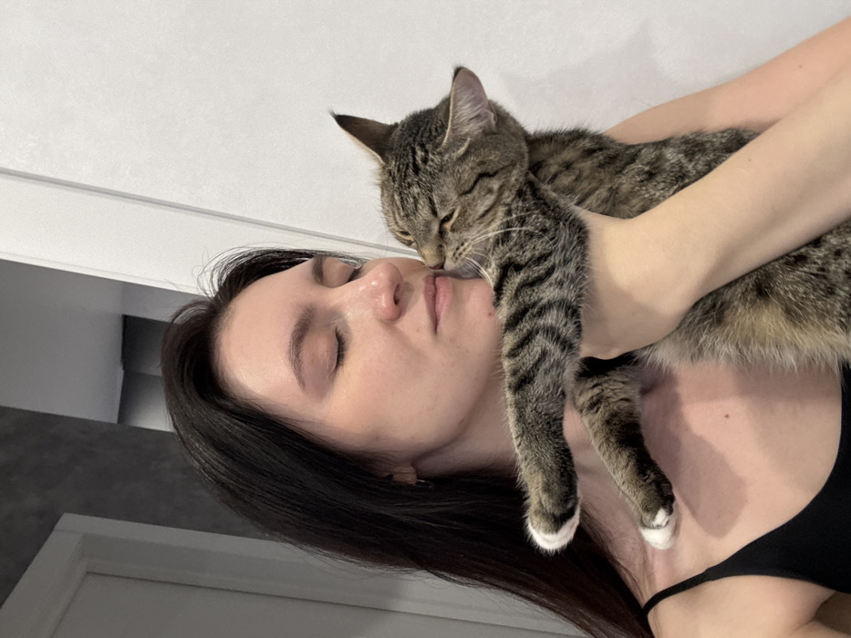 A female subject holds a tabby cat in an indoor domestic setting in Kazan, Russia. The woman, with long dark hair, wears a black tank top. Her eyes are closed, and her head is tilted towards the cat. The cat, exhibiting brown and black striped fur with white-tipped paws, is held by the woman's right arm, its face pressed against her right cheek/temple area, eyes also appearing closed. The background consists of a light-colored wall on the right and a white door frame on the left, with a darker grey wall segment visible through the door frame in the upper left.