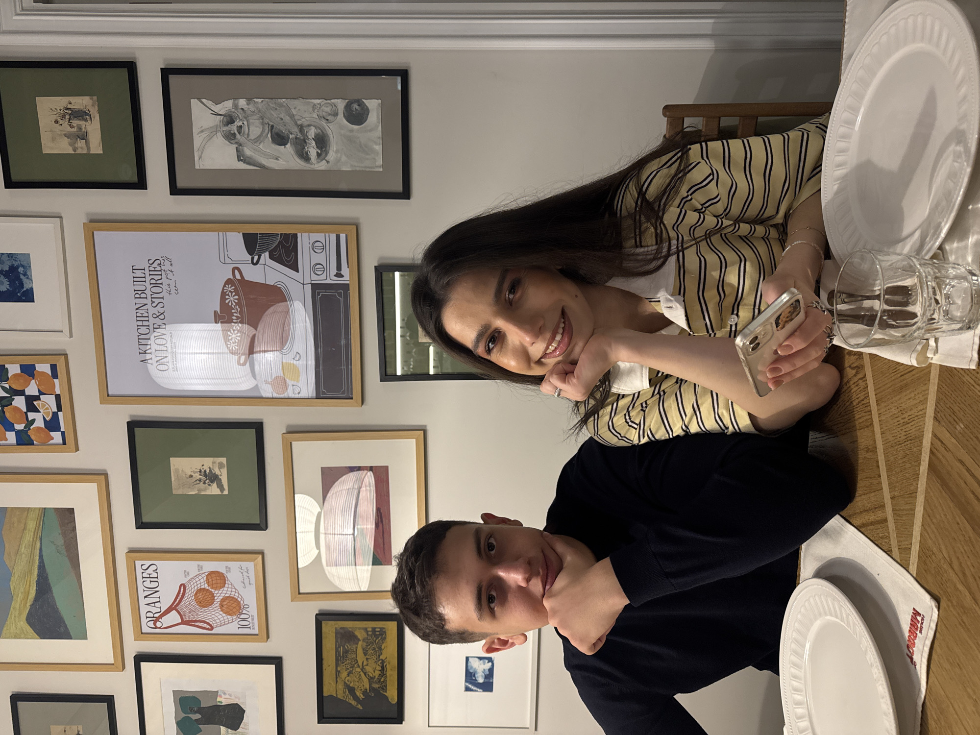 The image shows two individuals seated at a table, likely indoors, evidenced by the dining arrangement with plates, a glass, and cutlery. The setting is a room with a wall adorned with various framed artworks of different styles and sizes. The first individual has short, dark hair and is wearing a dark-colored shirt. The second individual, positioned behind the first, has long, dark hair and is wearing a striped shirt. One of the individuals is holding a cell phone. The environment appears domestic, possibly a dining room. The location within the city of Yerevan, Armenia, cannot be determined from the provided image.