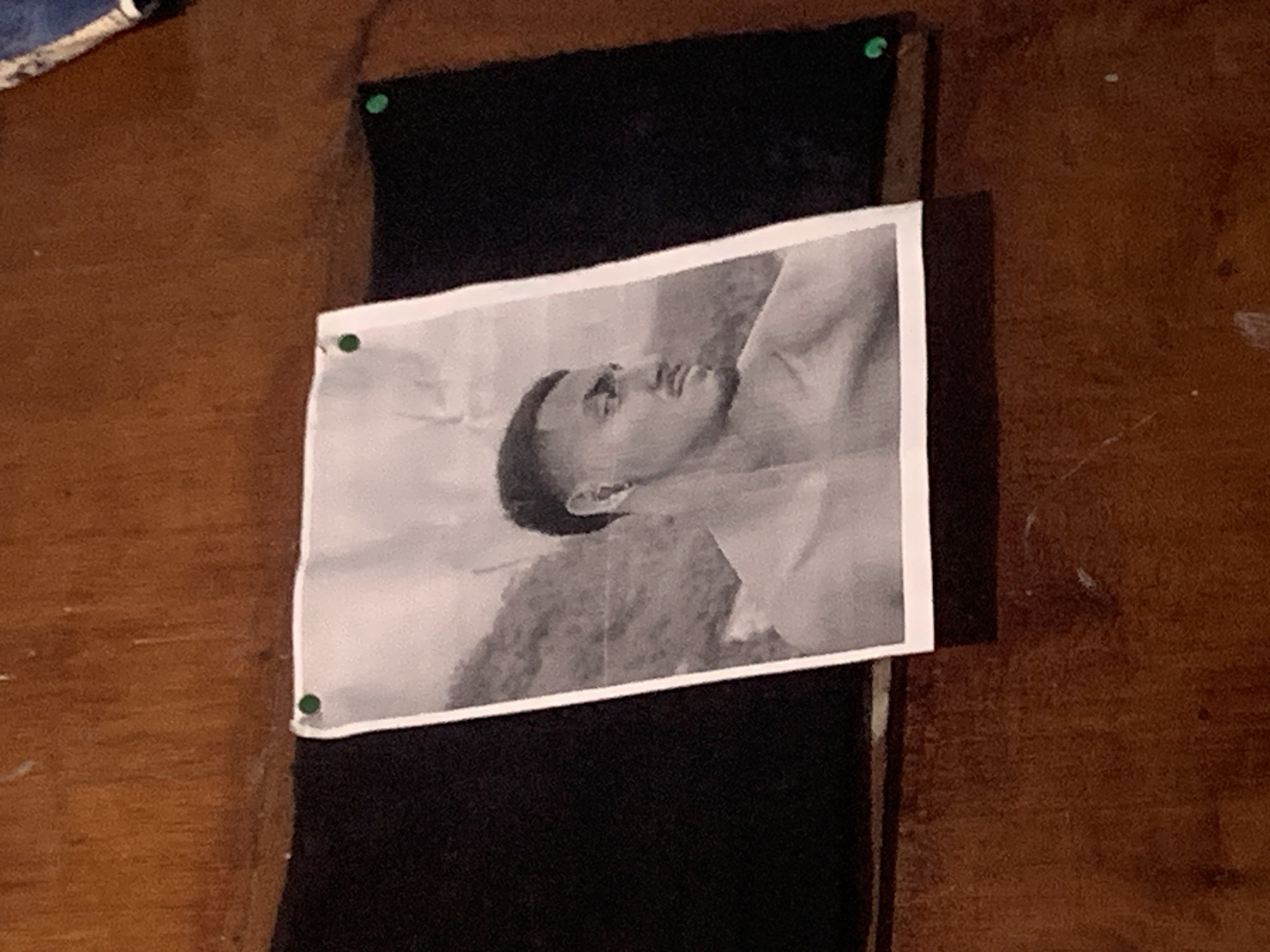 A black and white photograph of an individual is centrally displayed on a dark, vertical strip of fabric, which is pinned onto a reddish-brown wooden surface. The person in the photograph, likely a young male, is shown in profile, looking upwards with a calm expression. He has short, dark hair. The photograph is secured by four small, green thumbtacks, one in each corner. The dark fabric strip appears to be a plain, solid black textile. The wooden background features a visible grain and some subtle discoloration, suggesting an indoor setting like a wall or board. In the upper left corner of the frame, a small part of a blue and white patterned object is visible. The scene depicts a static display of a photograph, without any active human presence. There are no discernible details regarding the time of day or weather, nor any specific visual cues within the image itself to confirm the location as Santa Ana, Venezuela. No text is visible in the image.