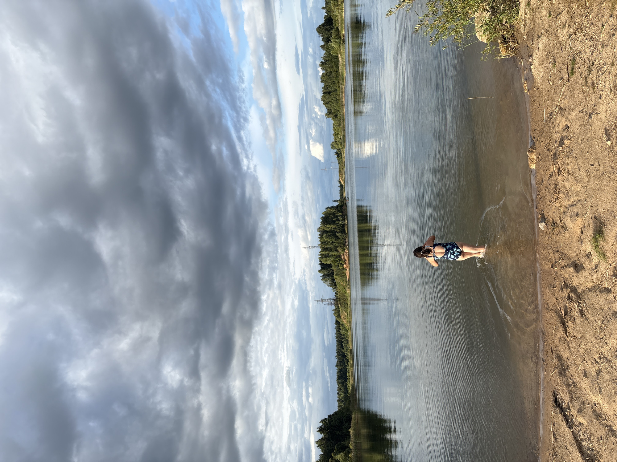 The image depicts a picturesque natural landscape with a calm body of water, likely a lake or a large pond, surrounded by lush green trees. A sandy shore is visible in the foreground. A young person, wearing a dark blue swimsuit with a floral pattern, is standing in the shallow water, facing away from the camera. They appear to be holding a mobile phone or camera in their hands, possibly taking a photograph or video. The sky is dramatic, filled with a mix of dark, heavy clouds and patches of blue sky, suggesting an overcast or impending weather change. In the background, on the far bank of the water, a dense forest stretches across the horizon, with several tall, industrial-looking towers visible above the treeline. The overall scene conveys a sense of tranquility and natural beauty, interrupted by the presence of man-made structures. The location is identified as being in the city of Zamost'e, Russia.